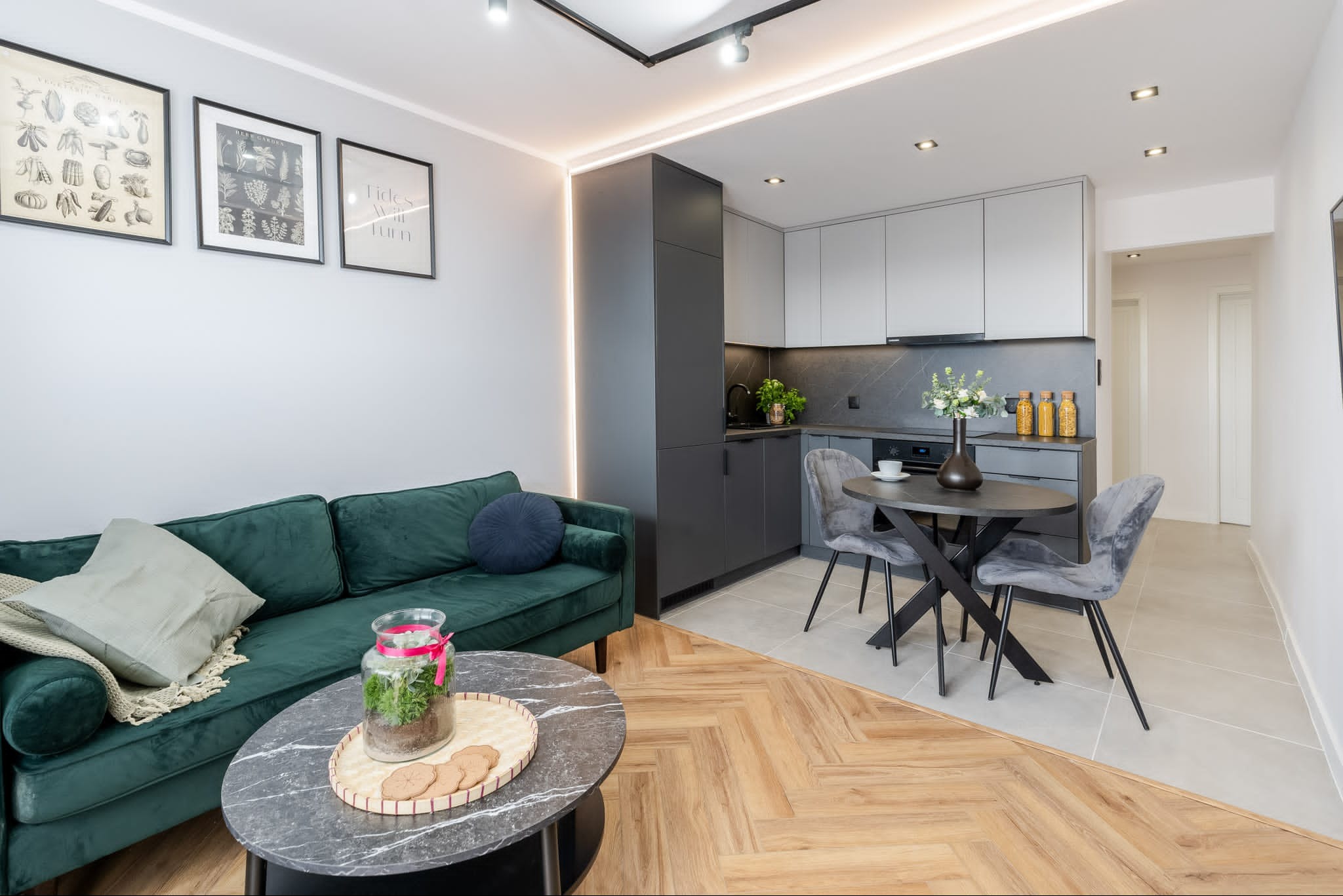 Nowoczesne wnętrze z aneksem kuchennym w odcieniach szarości, z zieloną sofą i okrągłym stolikiem kawowym na pierwszym planie, drewniana podłoga w jodełkę przechodzi w jasne płytki.