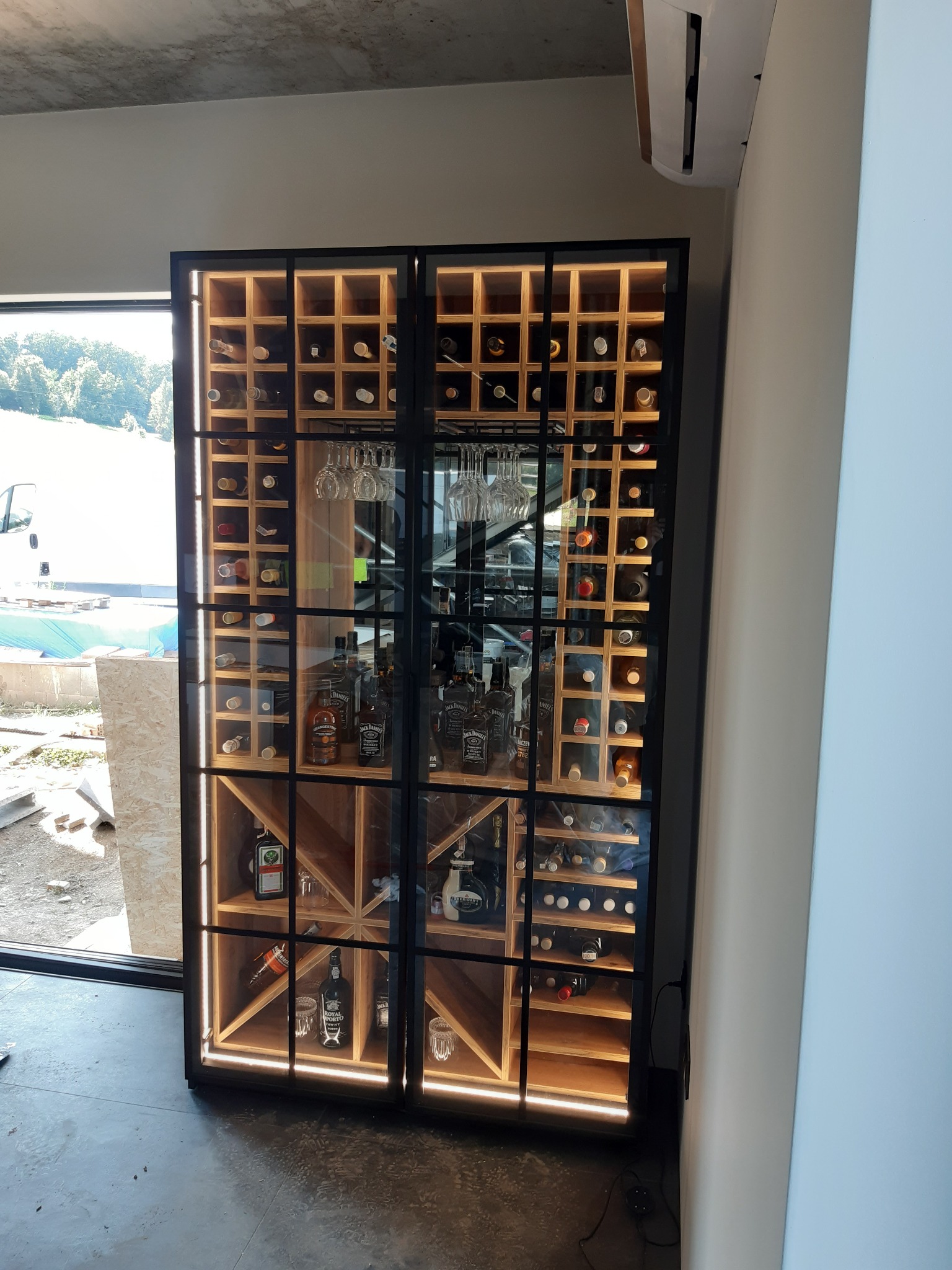 Duża, przeszklona szafa z drewnianymi półkami na wina i alkohole, podświetlana od wewnątrz, z widocznymi butelkami wina, whisky i kieliszkami, umieszczona w nowoczesnym wnętrzu z widokiem...