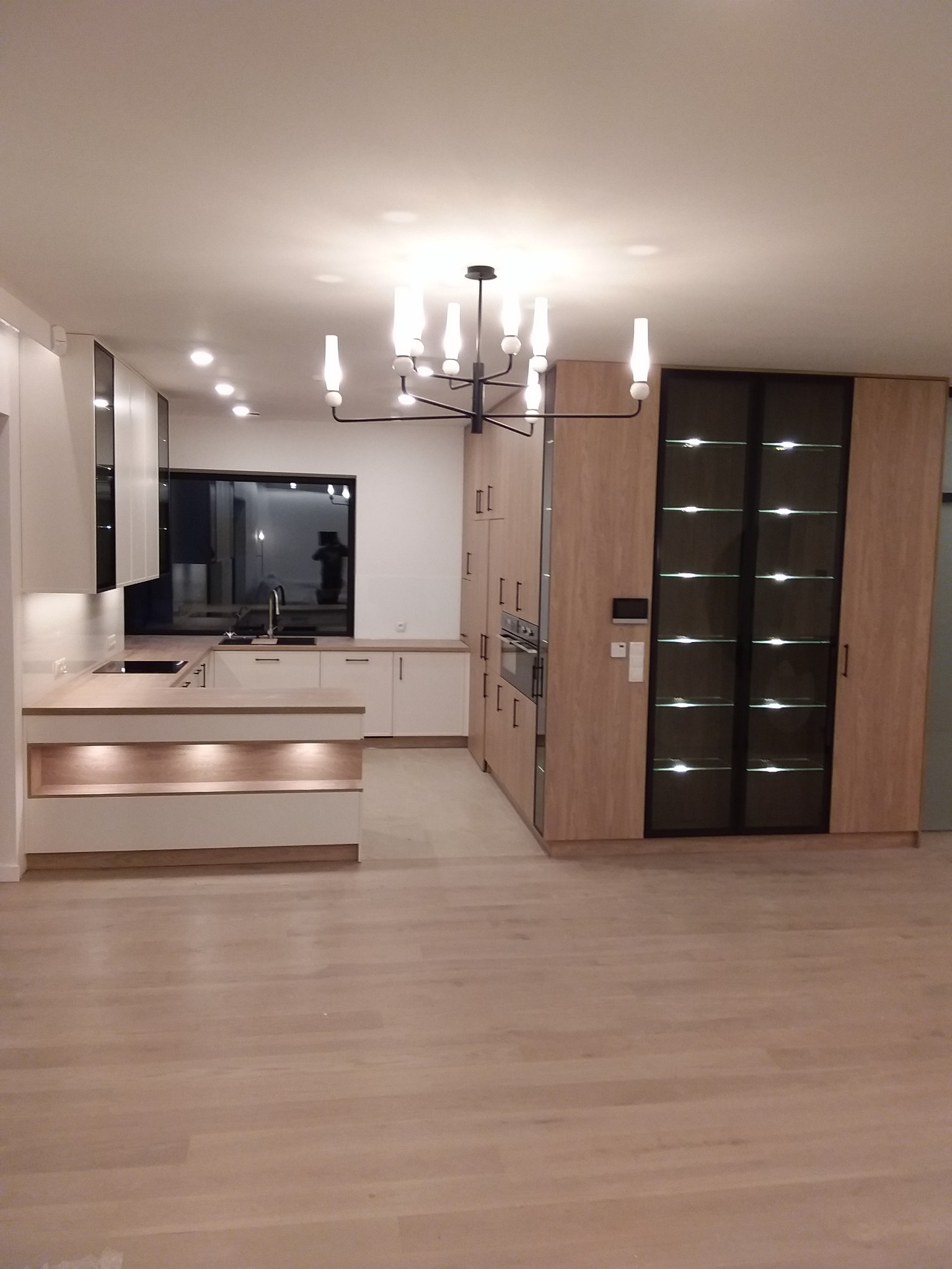 Nowoczesna kuchnia z białymi frontami szafek, drewnianym blatem, oświetleniem LED pod szafkami i dużą, przeszkloną szafą z podświetleniem w tle, oświetlona lampą wiszącą w stylu industrialnym.