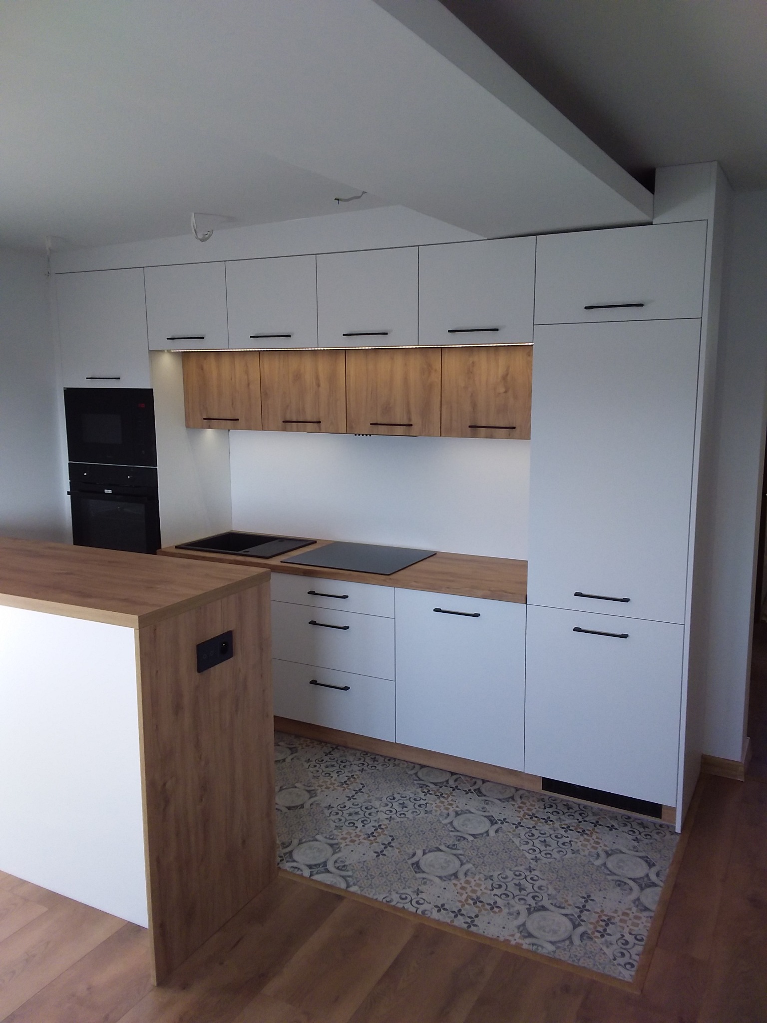 Nowoczesna kuchnia z białymi i drewnopodobnymi frontami, czarnymi uchwytami, płytą indukcyjną i piekarnikiem w zabudowie, oświetleniem podszafkowym LED i wzorzystą podłogą.