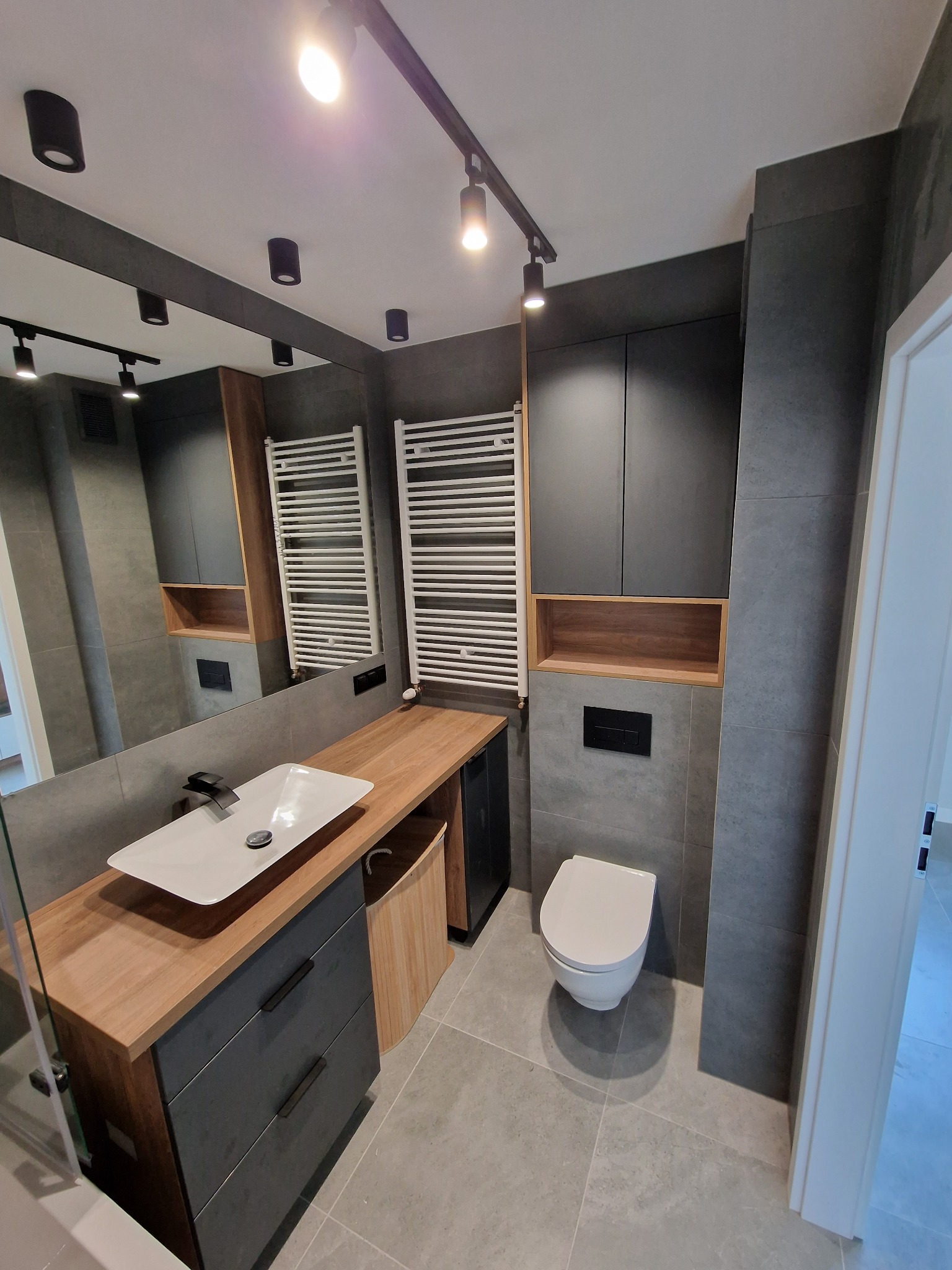 Nowoczesna łazienka z szarymi płytkami, drewnianym blatem pod umywalką, ciemnymi szafkami i podwieszaną toaletą, oświetlona punktowymi lampami na szynoprzewodzie.