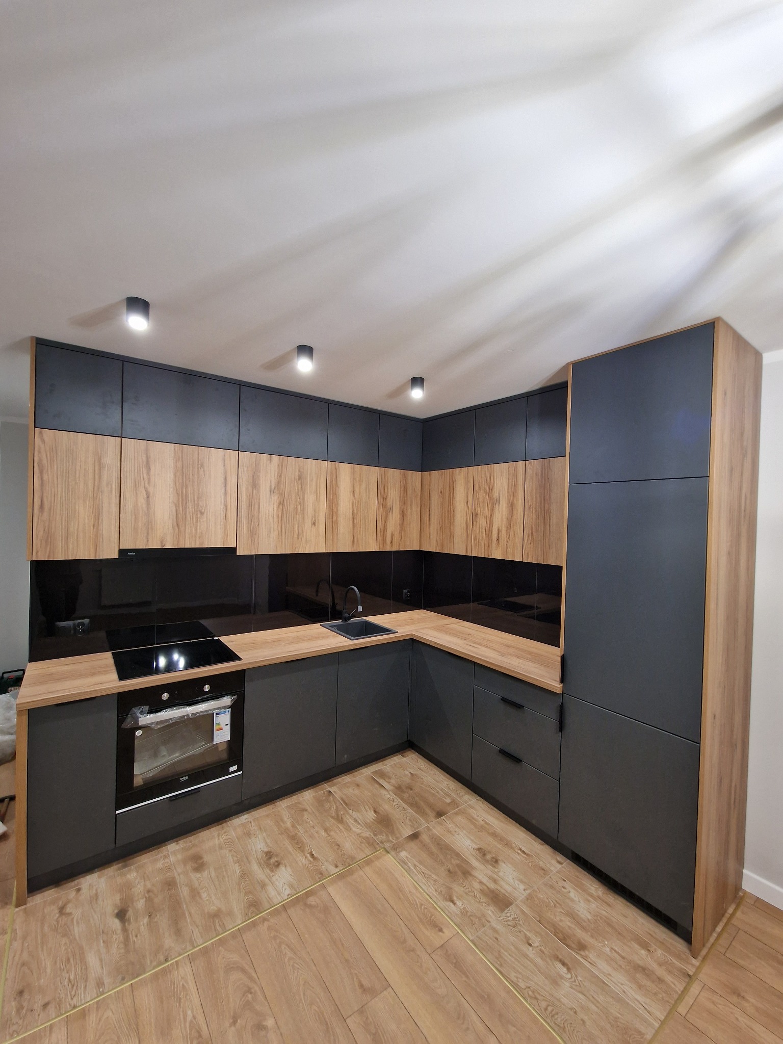 Nowoczesna kuchnia narożna z frontami w kolorze dębu i grafitu, czarny szklany panel nad blatem, drewnopodobna podłoga, piekarnik z widoczną etykietą energetyczną.