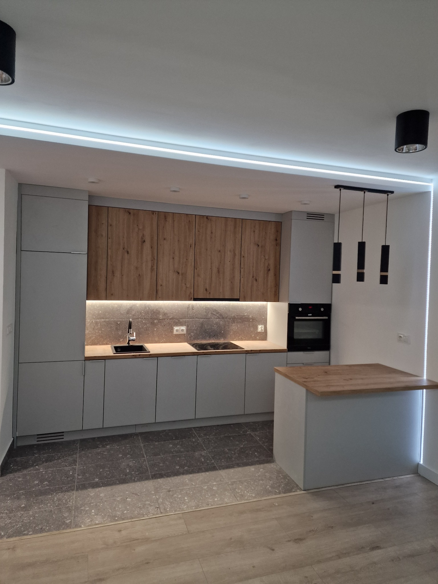 Nowoczesna kuchnia z szarymi szafkami dolnymi i górnymi z drewnianymi frontami, podświetleniem LED pod szafkami górnymi, czarnym zlewem i płytą, oraz wyspą kuchenną z drewnianym blatem i trzema...