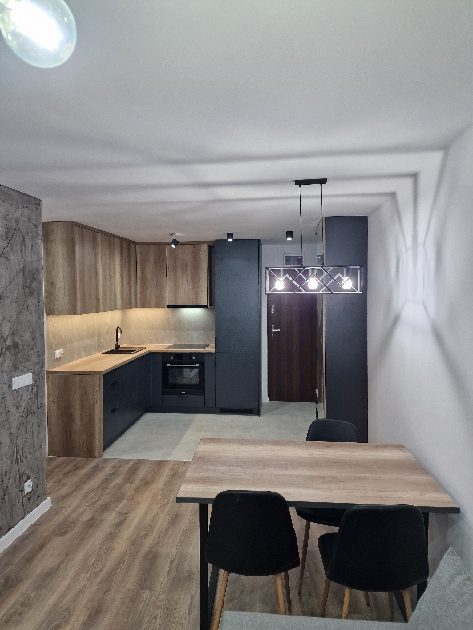 Nowoczesna kuchnia z drewnianymi szafkami górnymi i granatowymi dolnymi, jasny blat, czarny zlew i armatura, stół z czarnymi krzesłami, designerska lampa wisząca z trzema żarówkami, ściana...