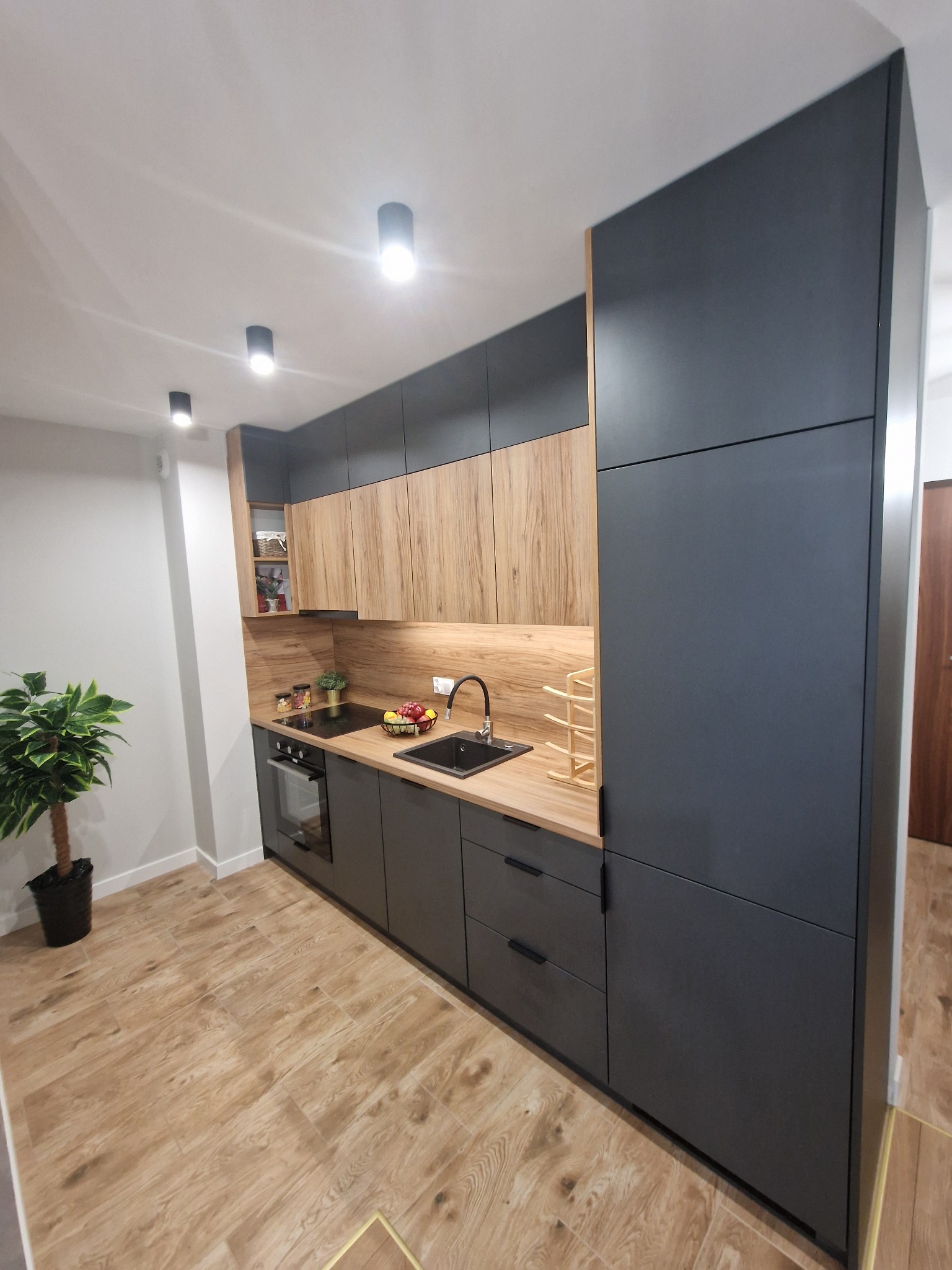 Nowoczesna kuchnia z ciemnoszarymi szafkami dolnymi i drewnopodobnymi szafkami górnymi, czarnym zlewem i blatem z jasnego drewna, oświetlona punktowymi lampami sufitowymi, z rośliną doniczkową w rogu.
