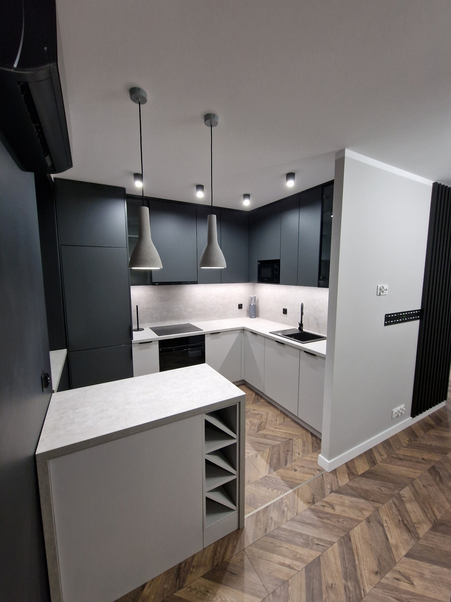 Nowoczesna kuchnia z ciemnymi, matowymi frontami szafek górnych i jasnymi dolnymi, z wyspą wyposażoną w półki na wino i oświetleniem punktowym oraz wiszącymi lampami w kształcie stożków,...