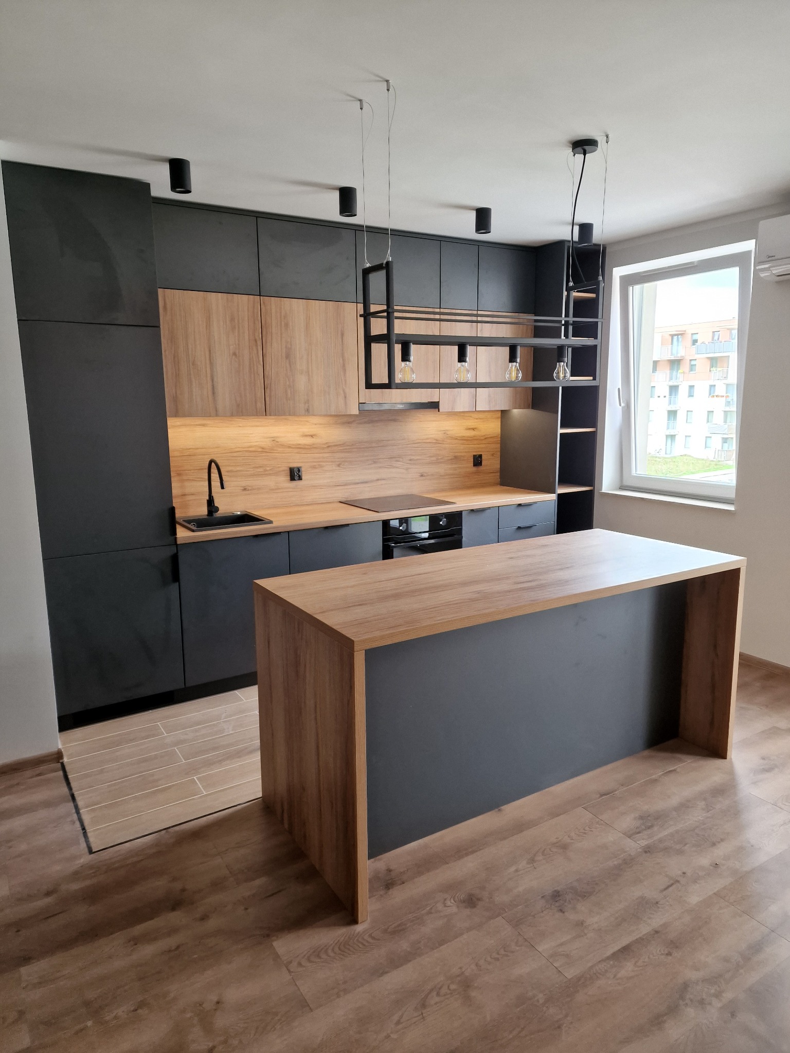 Nowoczesna kuchnia z wyspą, fronty szafek w kolorze grafitowym i drewnopodobnym, czarna armatura i oświetlenie, widok na okno.