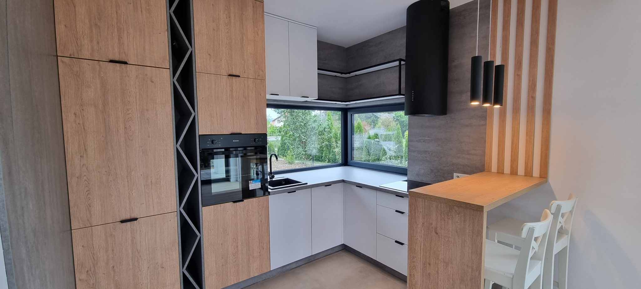 Nowoczesna kuchnia z drewnianymi i białymi frontami, czarnym okapem cylindrycznym i barem z białymi krzesłami.