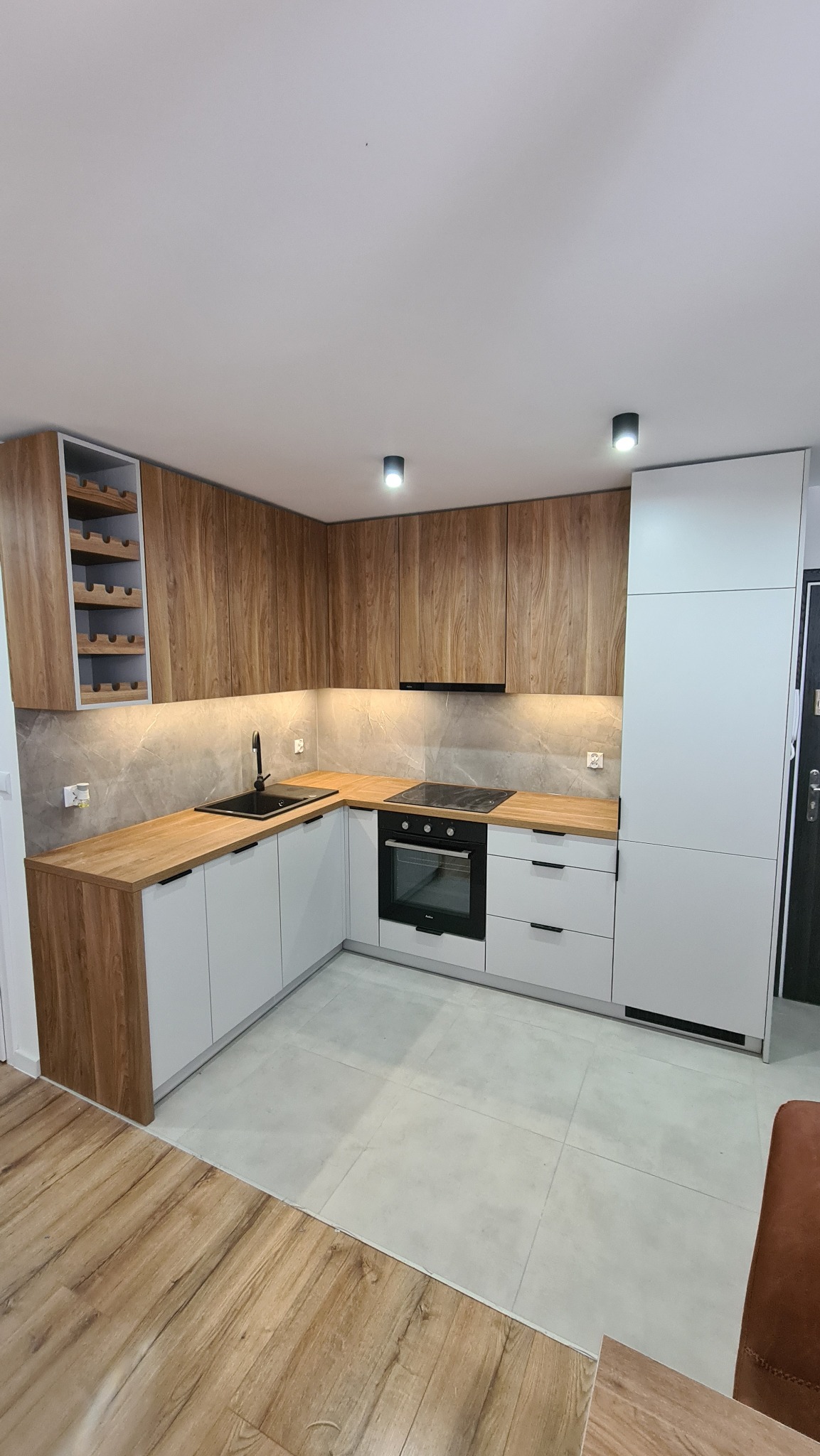 Nowoczesna kuchnia w kształcie litery L z drewnianymi frontami górnych szafek i blatem, białymi dolnymi szafkami i szarą mozaiką na ścianie, oświetlona punktowymi lampami sufitowymi i listwą LED...