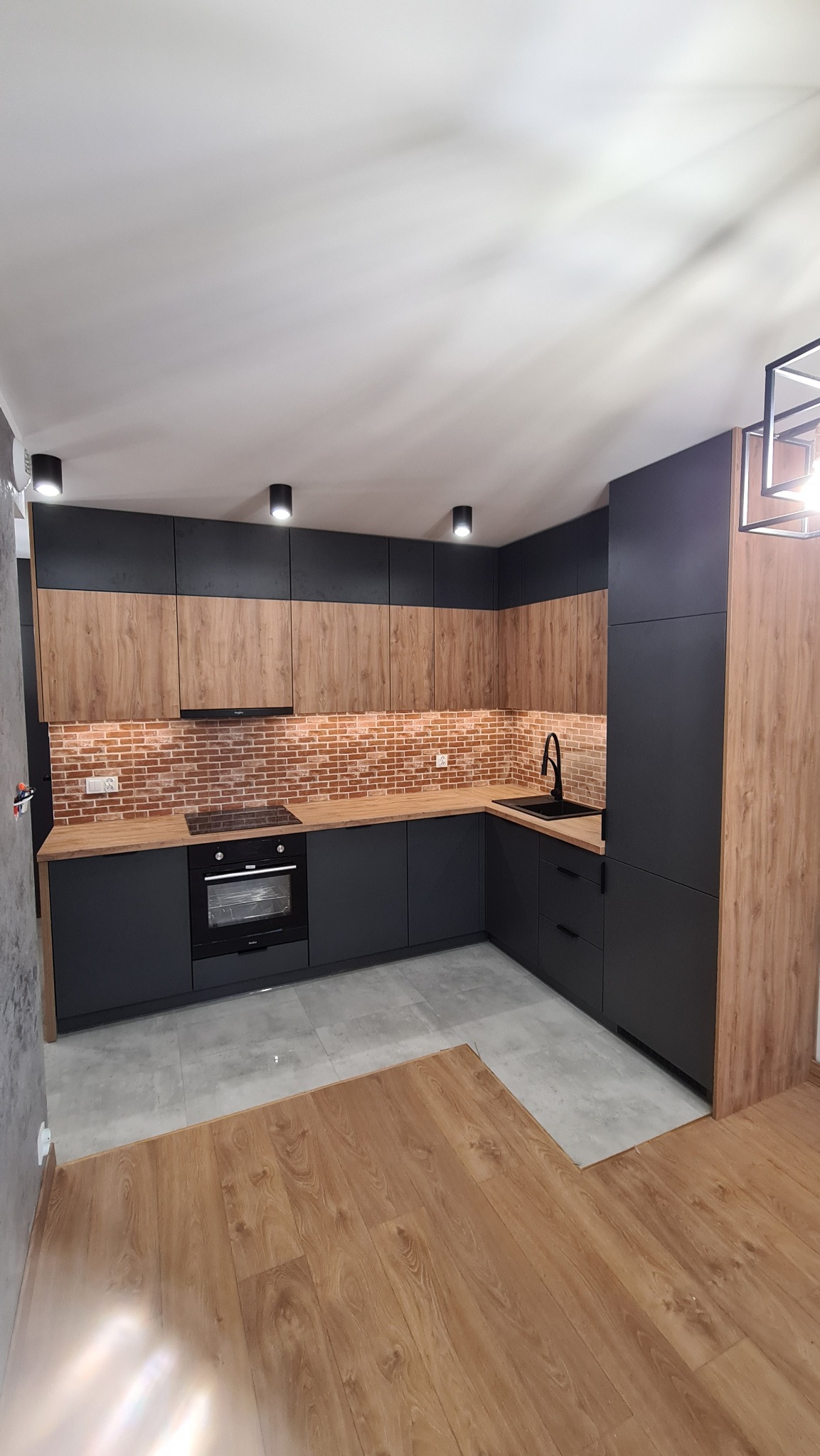 Nowoczesna kuchnia z czarnymi dolnymi szafkami i drewnopodobnymi górnymi, ceglana ściana nad blatem, czarny zlewozmywak i bateria, oświetlenie punktowe, fragment drewnianej podłogi.