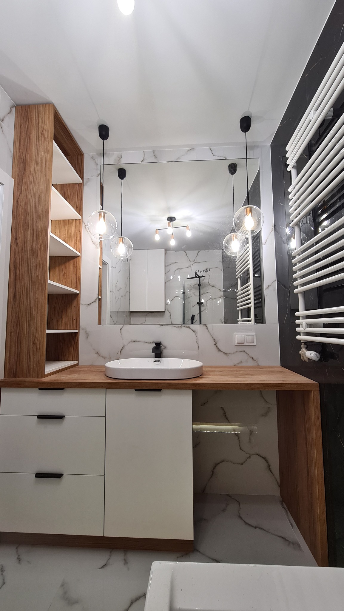 Nowoczesna łazienka z białą umywalką nablatową, drewnianym blatem, szafkami z minimalistycznymi czarnymi uchwytami, marmurowymi płytkami na ścianach i podłodze oraz dekoracyjnym oświetleniem.