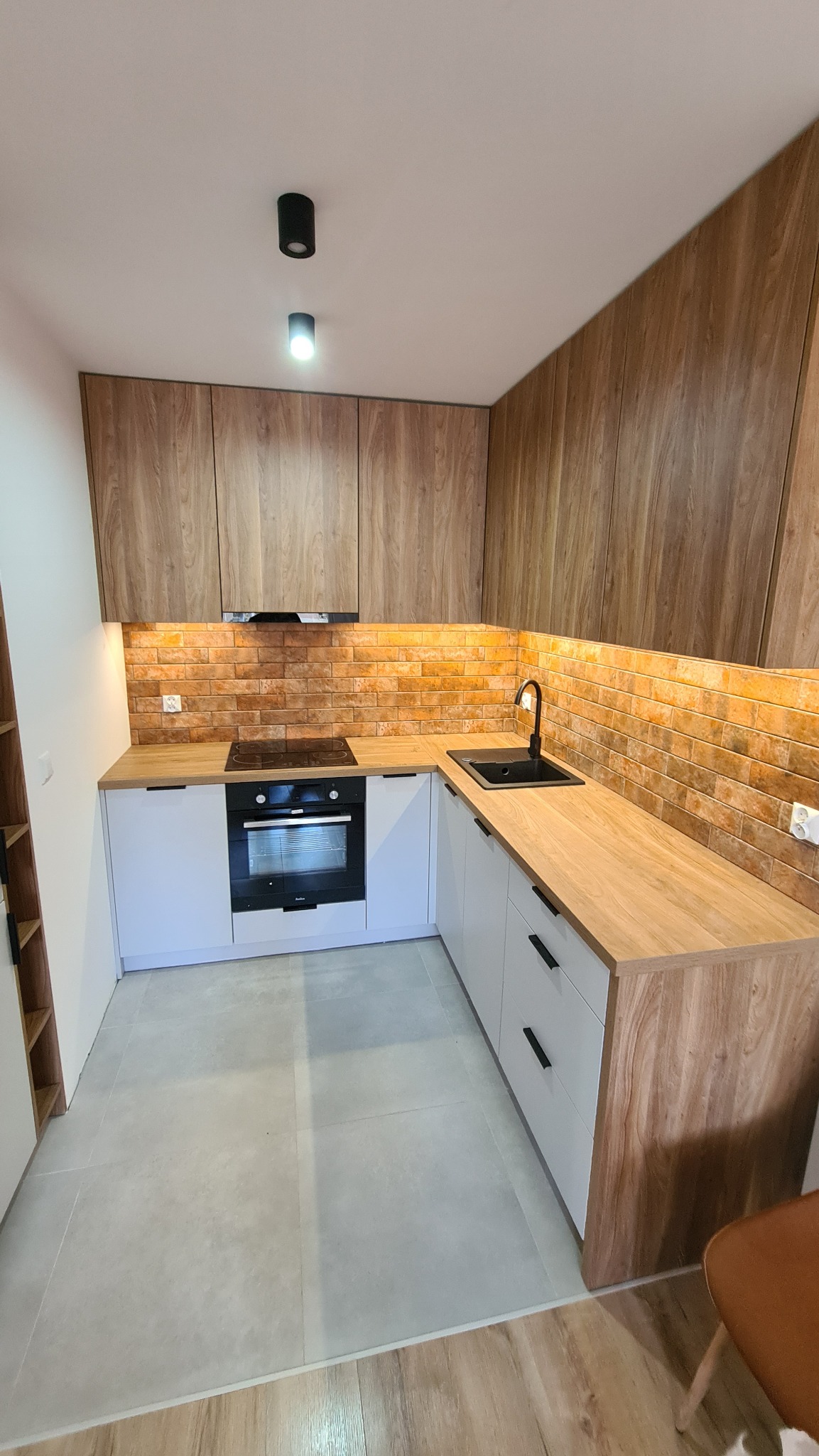 Kompaktowa kuchnia w kształcie litery L z białymi szafkami dolnymi i górnymi w odcieniach drewna, ceglana ściana nad blatem, czarny zlew i armatura, oświetlenie podszafkowe i punktowe.