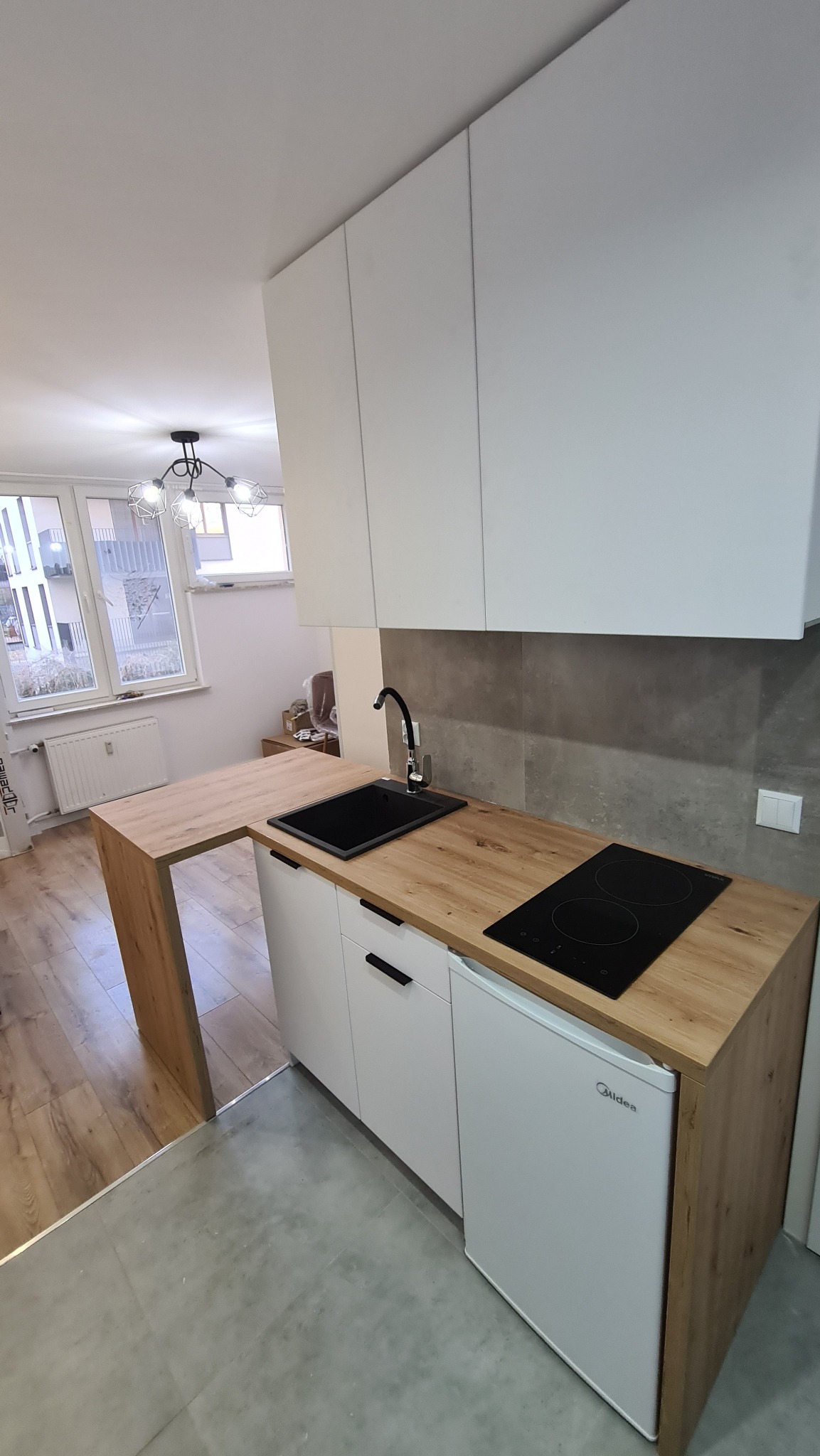 Nowoczesna, minimalistyczna kuchnia z białymi szafkami i blatem imitującym drewno, z czarnym zlewem i płytą indukcyjną, oraz małą lodówką w zabudowie.