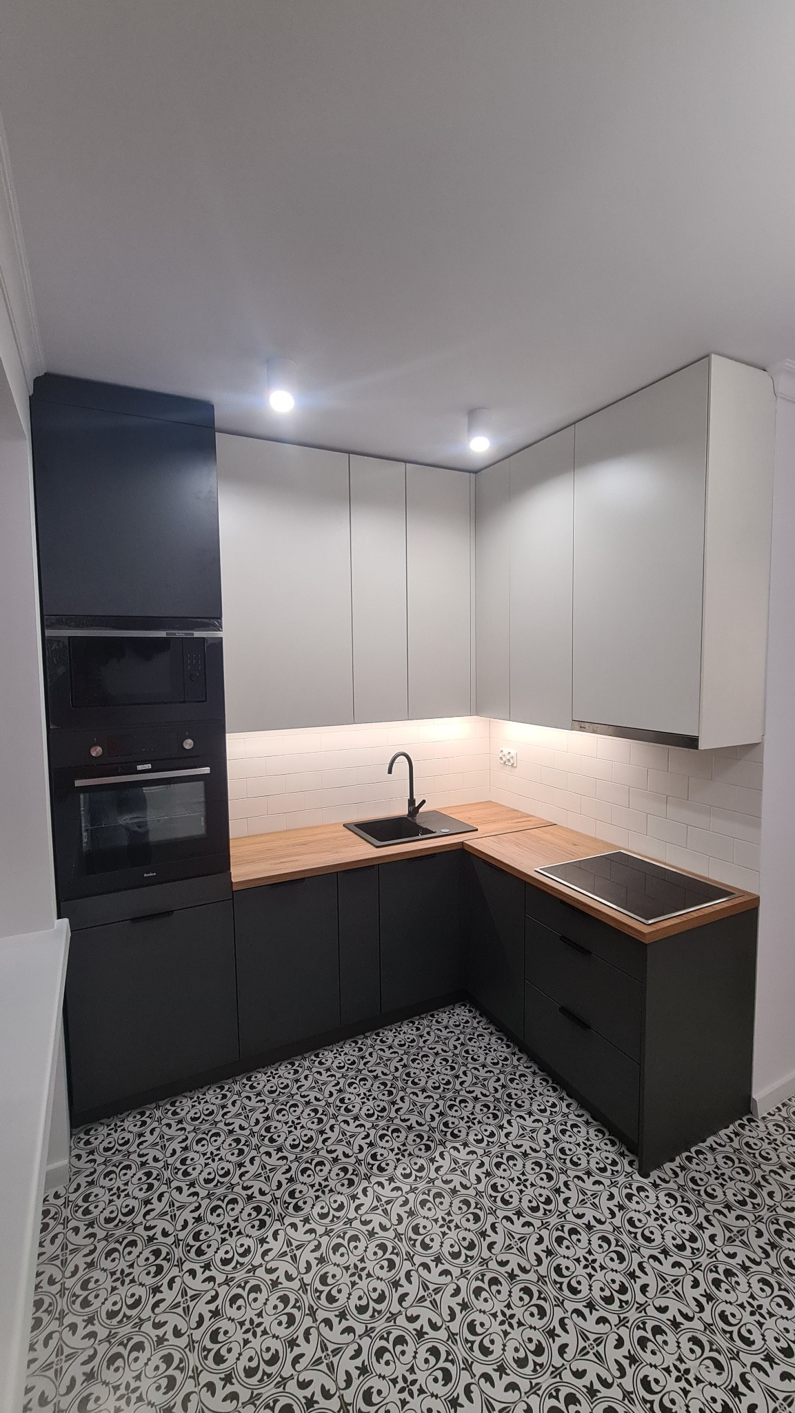 Nowoczesna kuchnia w kształcie litery L z czarnymi dolnymi szafkami, drewnianym blatem, białymi górnymi szafkami i wzorzystą podłogą.