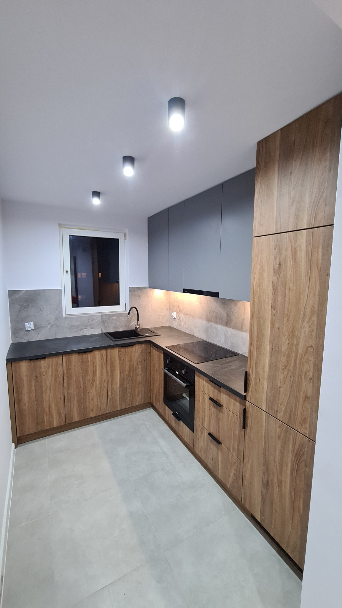 Nowoczesna kuchnia z drewnianymi frontami szafek dolnych i szarymi górnymi, czarny zlew i bateria, szary blat oraz marmurowe płytki na ścianie, oświetlenie punktowe sufitowe i podszafkowe.