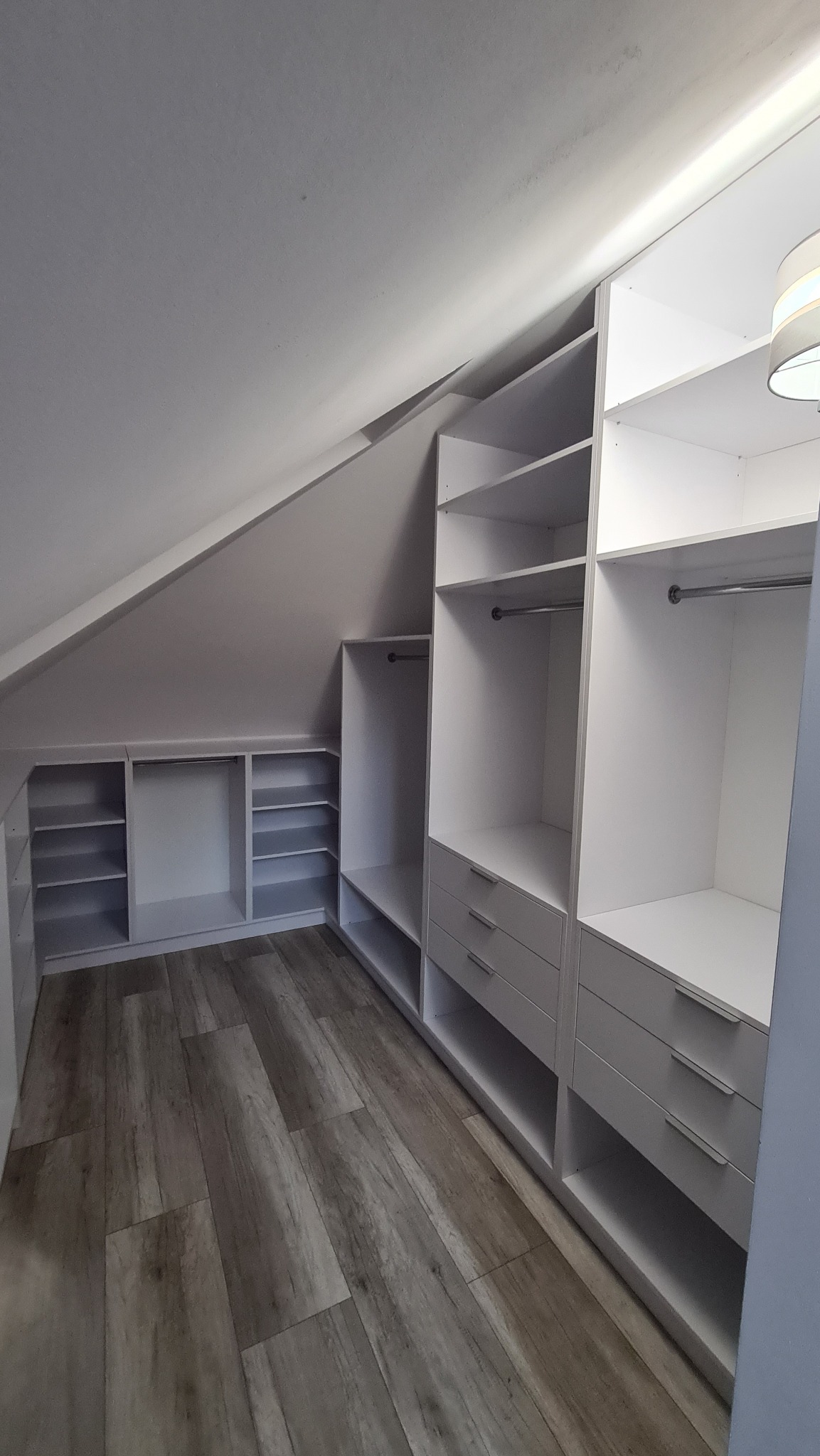 Biała zabudowa garderoby na poddaszu ze skośnym sufitem, widoczne półki, szuflady i drążki na ubrania, podłoga z paneli imitujących drewno.