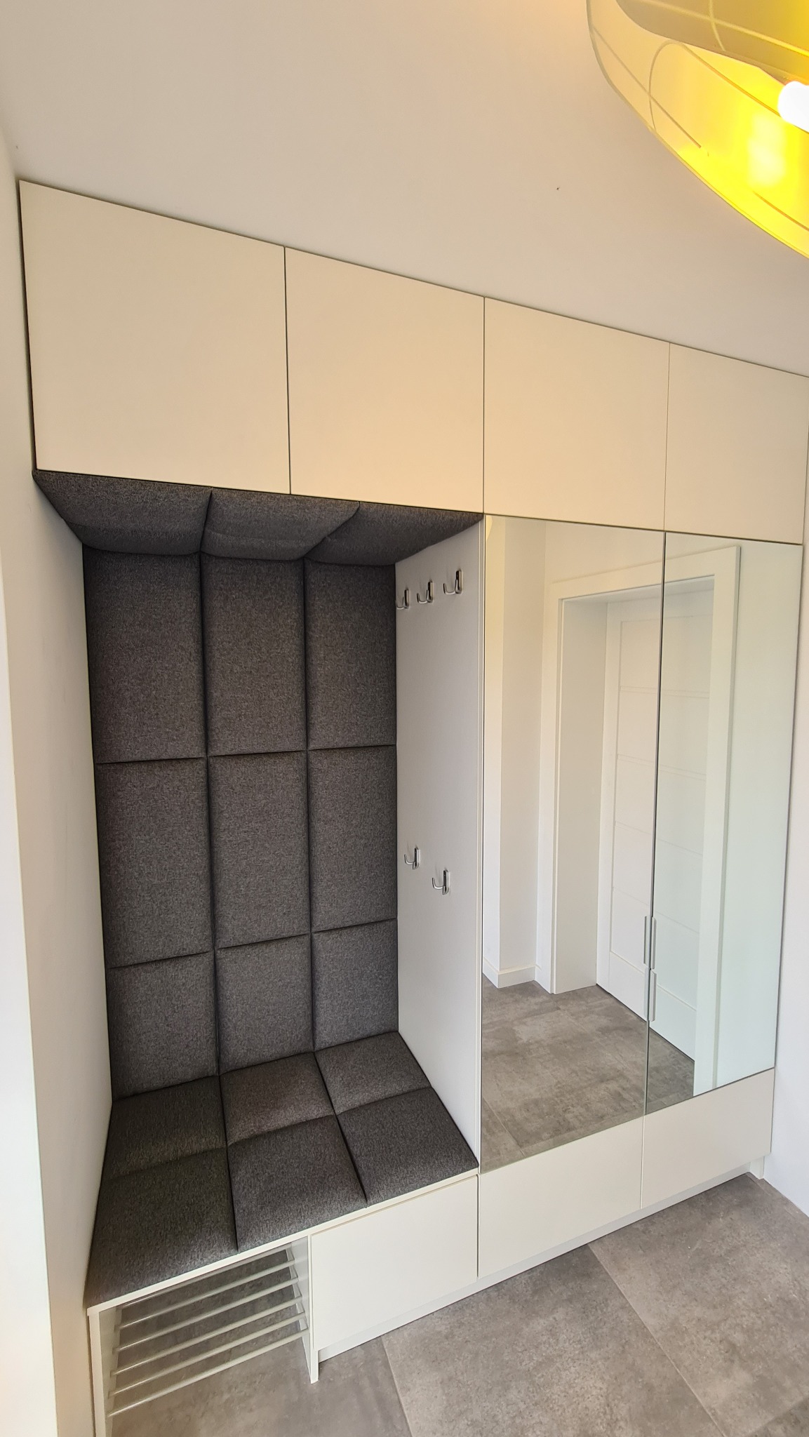 Biała zabudowa przedpokoju z siedziskiem obitym szarą tkaniną w kwadraty, wbudowane lustro oraz półka na buty.