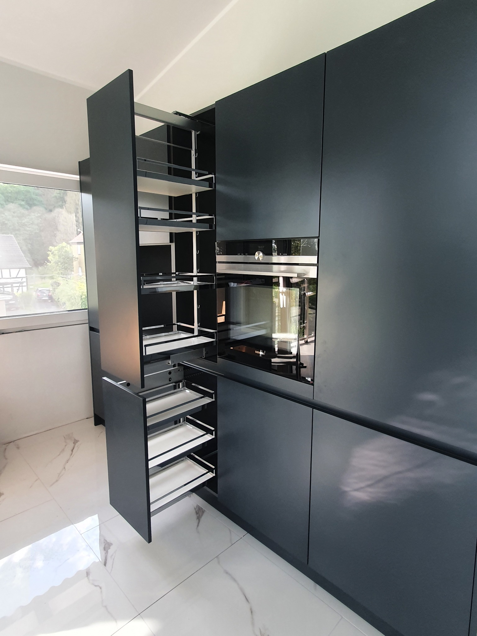 Nowoczesna, grafitowa zabudowa kuchenna z wysuwanymi półkami cargo i piekarnikiem w zabudowie, widok na marmurowe płytki podłogowe i okno.