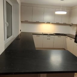 Zakład Kamieniarski Wojciech Lociński - Nowoczesna kuchnia z czarnym, kamiennym blatem w kształcie litery L, jasnymi szafkami i oknem po lewej stronie, oświetlona wiszącą lampą.