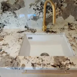 Biały zlew wmontowany w blat kuchenny z kamienia naturalnego o wzorze przypominającym chmury, bateria w kolorze szczotkowanego złota, złoty uchwyt szafki.