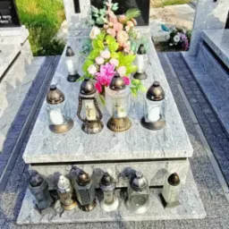 Granitowy pomnik z czarną tablicą, ozdobiony kwiatami i lampionami, otoczony szarą kostką brukową.