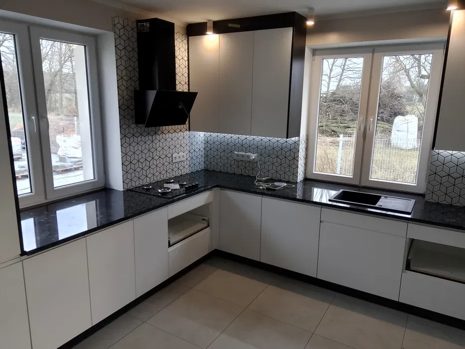 Nowoczesna kuchnia z czarnym blatem i białymi szafkami, widoczne okno z widokiem na zewnątrz, płytki w geometryczny wzór na ścianie.