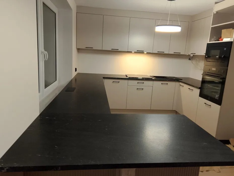 Nowoczesna kuchnia z czarnym, kamiennym blatem w kształcie litery L, jasnymi szafkami i oknem po lewej stronie, oświetlona wiszącą lampą.