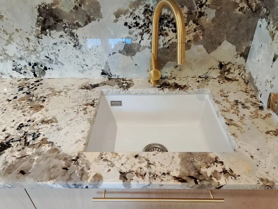 Biały zlew wmontowany w blat kuchenny z kamienia naturalnego o wzorze przypominającym chmury, bateria w kolorze szczotkowanego złota, złoty uchwyt szafki.