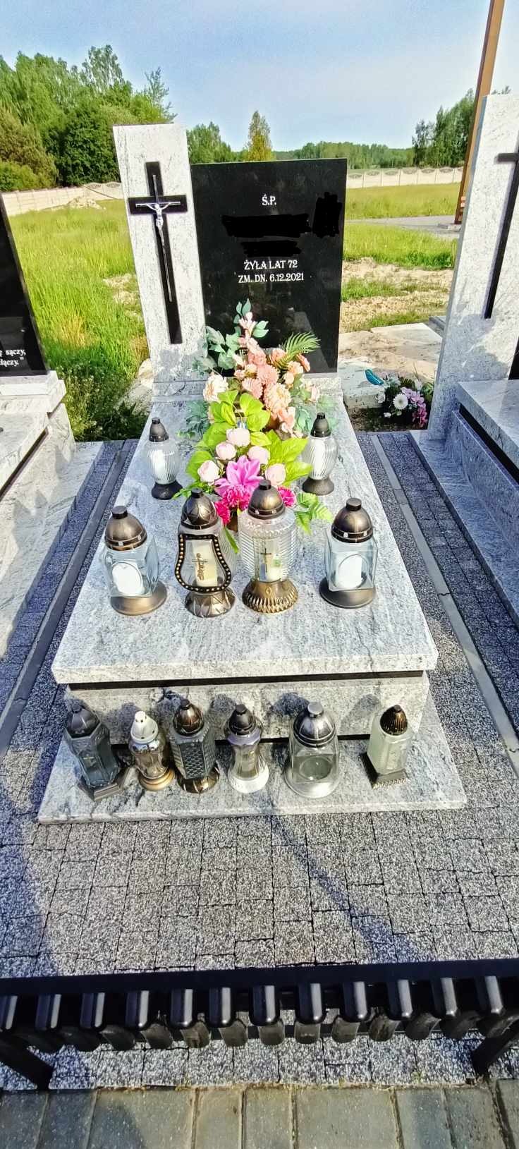 Granitowy pomnik z czarną tablicą, ozdobiony kwiatami i lampionami, otoczony szarą kostką brukową.