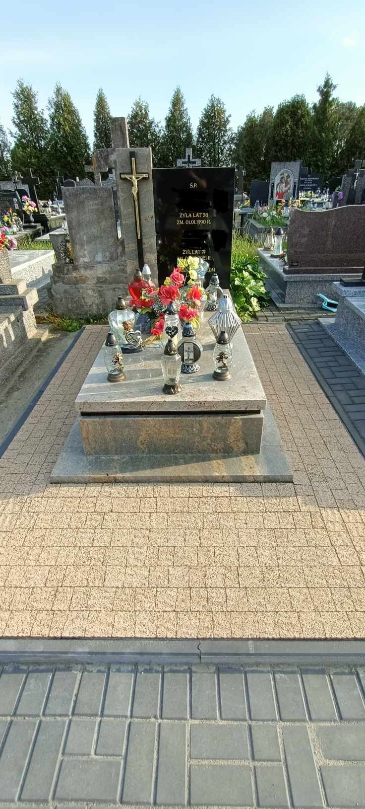 Granitowy pomnik z czarną płytą nagrobkową, ozdobiony kwiatami i lampionami, otoczony kostką brukową w odcieniach beżu i szarości na cmentarzu.