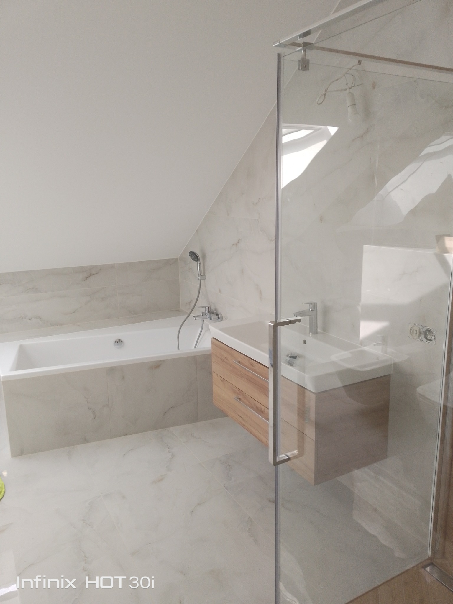 Jasna łazienka na poddaszu z wanną, umywalką z szafką z drewnianymi szufladami i kabiną prysznicową. Marmurowe płytki na ścianach i podłodze.