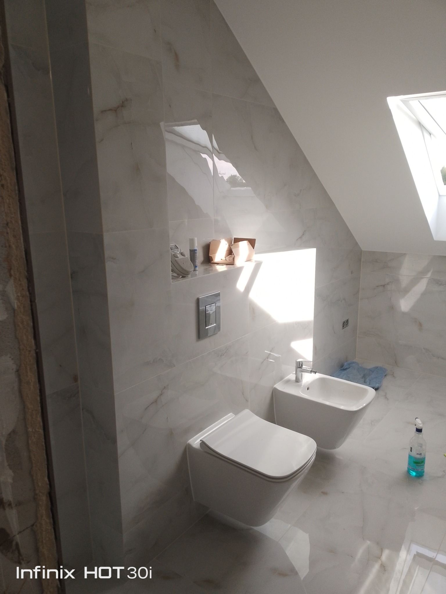 Wykończona łazienka na poddaszu z marmurowymi płytkami, wiszącą toaletą i bidetem. W tle okno dachowe i półka z akcesoriami. Widoczny fragment niedokończonej ściany.