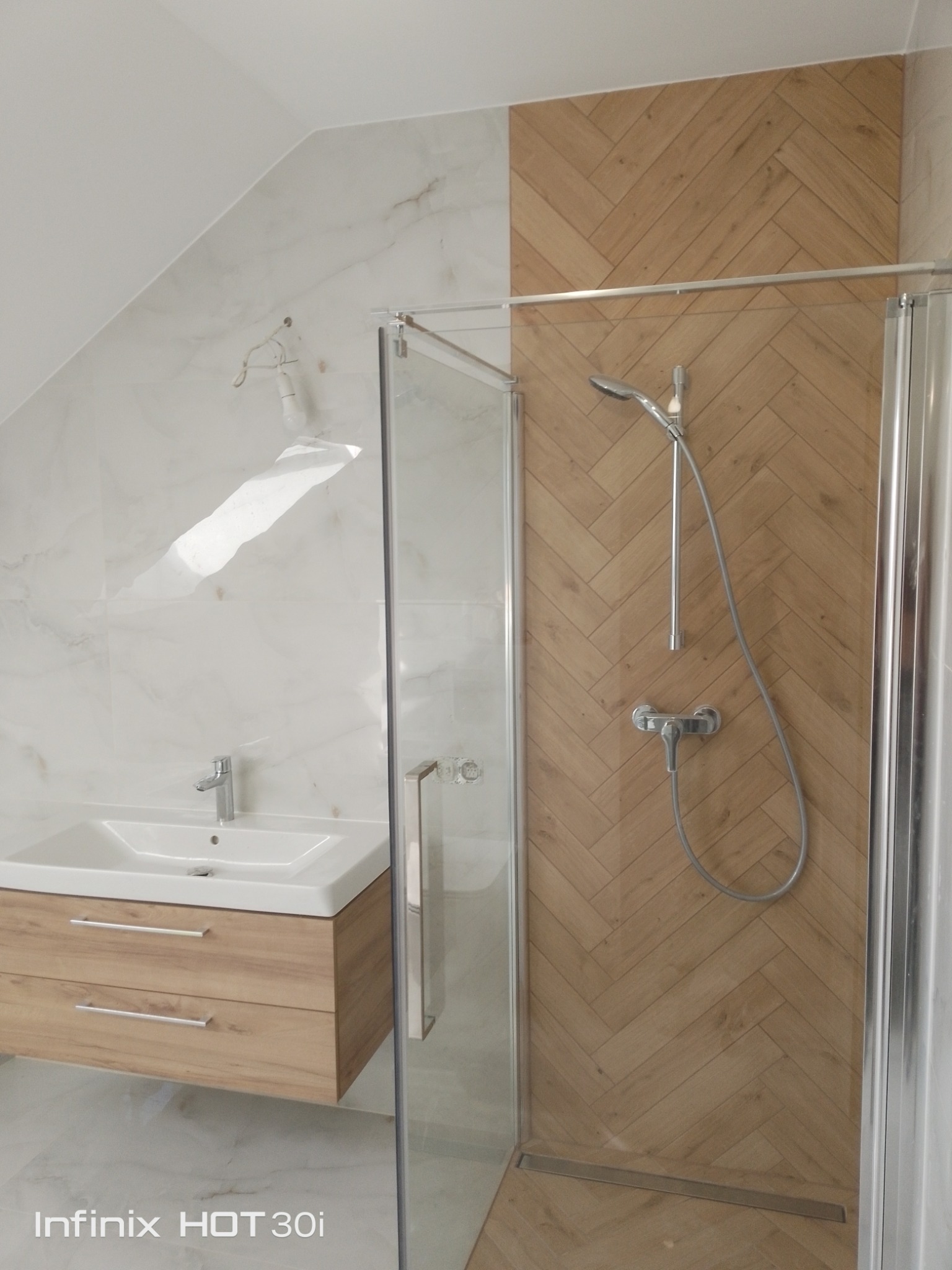 Nowoczesna łazienka na poddaszu z kabiną prysznicową, umywalką z szafką w odcieniach drewna i marmurowymi płytkami. Ukośny sufit dodaje unikalnego charakteru.