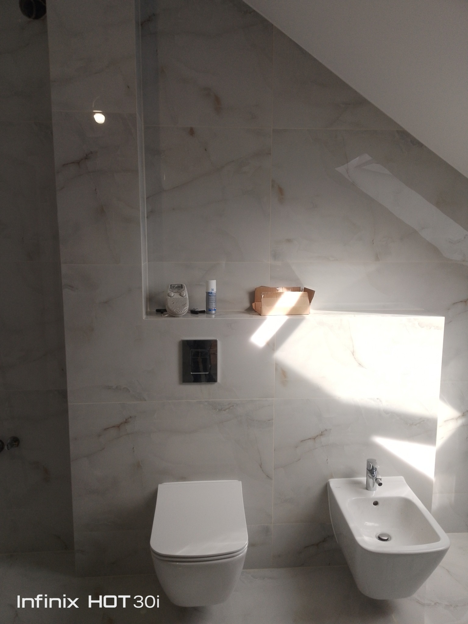 Wykończona łazienka z marmurowymi płytkami, wiszącą toaletą i bidetem. Na ścianie wnęka z dekoracjami i włącznik światła. Ukośny sufit. Promienie światła padają na płytki.