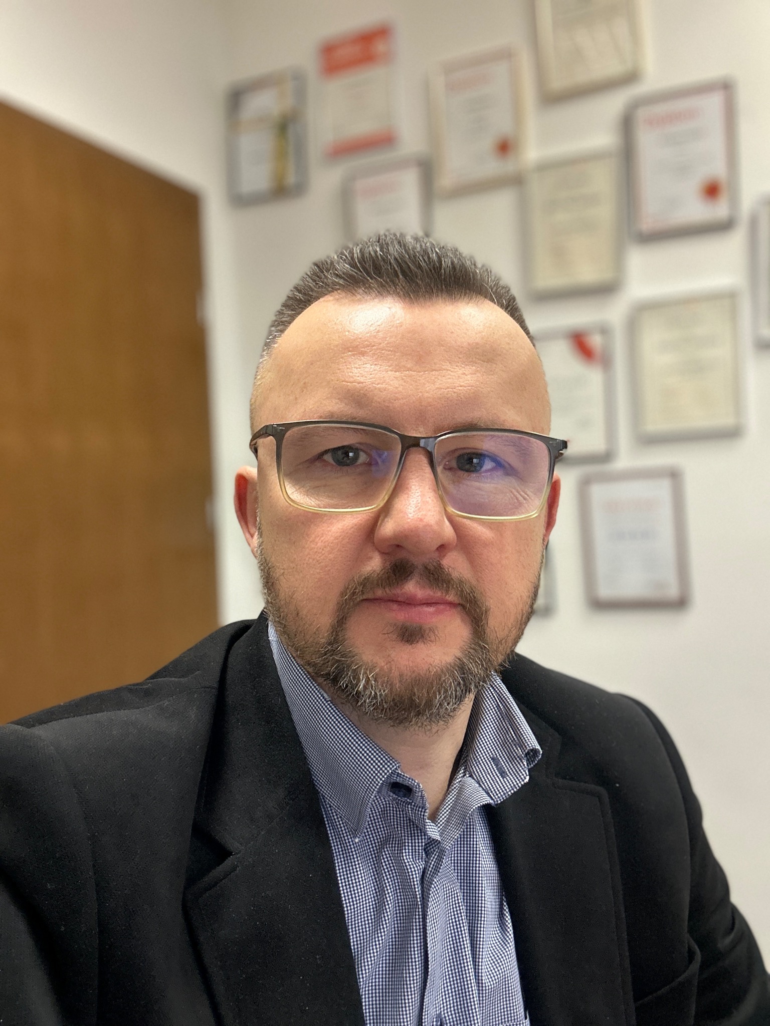 Portret mężczyzny w okularach i marynarce, w tle ściana z dyplomami i brązowe drzwi