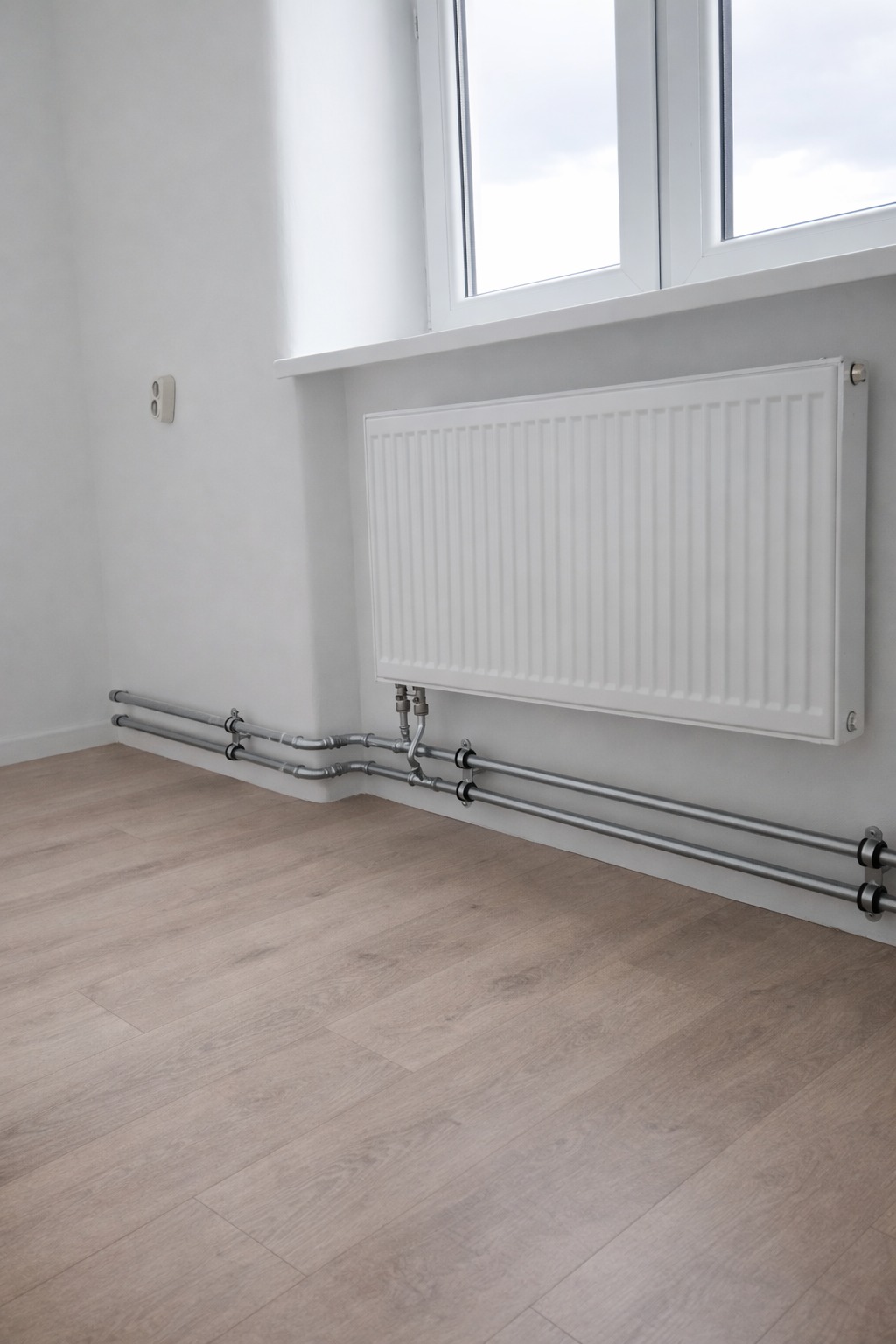Biały grzejnik panelowy zamontowany pod oknem, z widocznymi rurami doprowadzającymi, na tle jasnej ściany i podłogi z paneli. Minimalistyczny design.