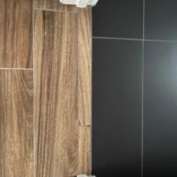 Połączenie płytek: imitacja drewna i czarne, kwadratowe. Widoczne fugi i listwa przypodłogowa. Detal wykończenia podłogi, bliski kadr.