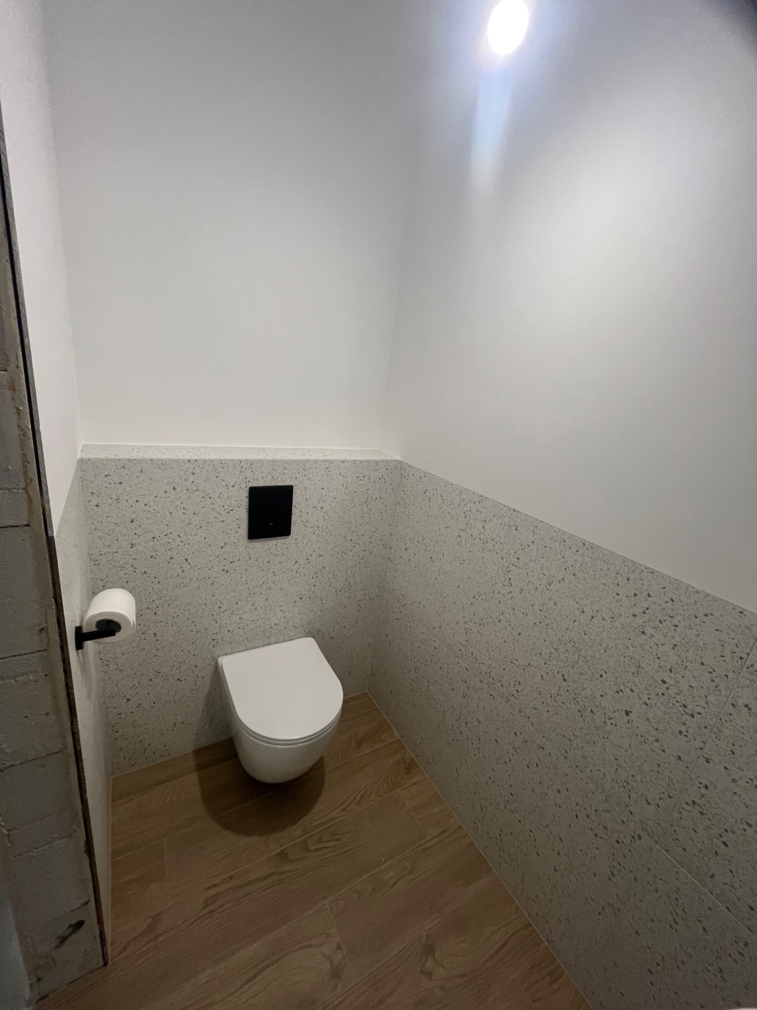 Nowoczesna, minimalistyczna łazienka z drewnopodobną podłogą, mozaiką na ścianach do połowy wysokości i podwieszaną toaletą z czarnym przyciskiem.