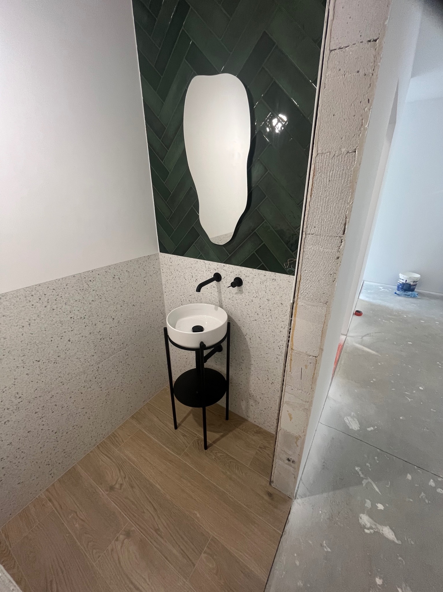 Nowoczesna łazienka z zieloną glazurą w jodełkę, asymetrycznym lustrem i wolnostojącą umywalką na tle surowego muru w trakcie wykończenia.