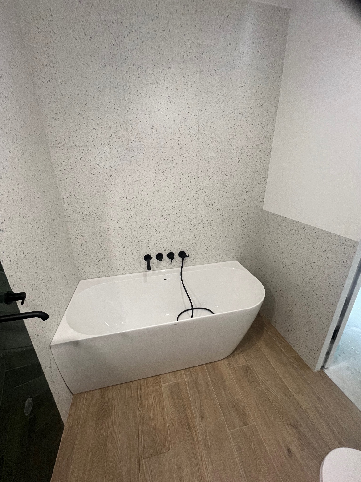 Nowoczesna łazienka z białą wanną, czarną armaturą i ścianami w jasny wzór lastryko. Podłoga wyłożona panelami imitującymi drewno. Minimalistyczny design.