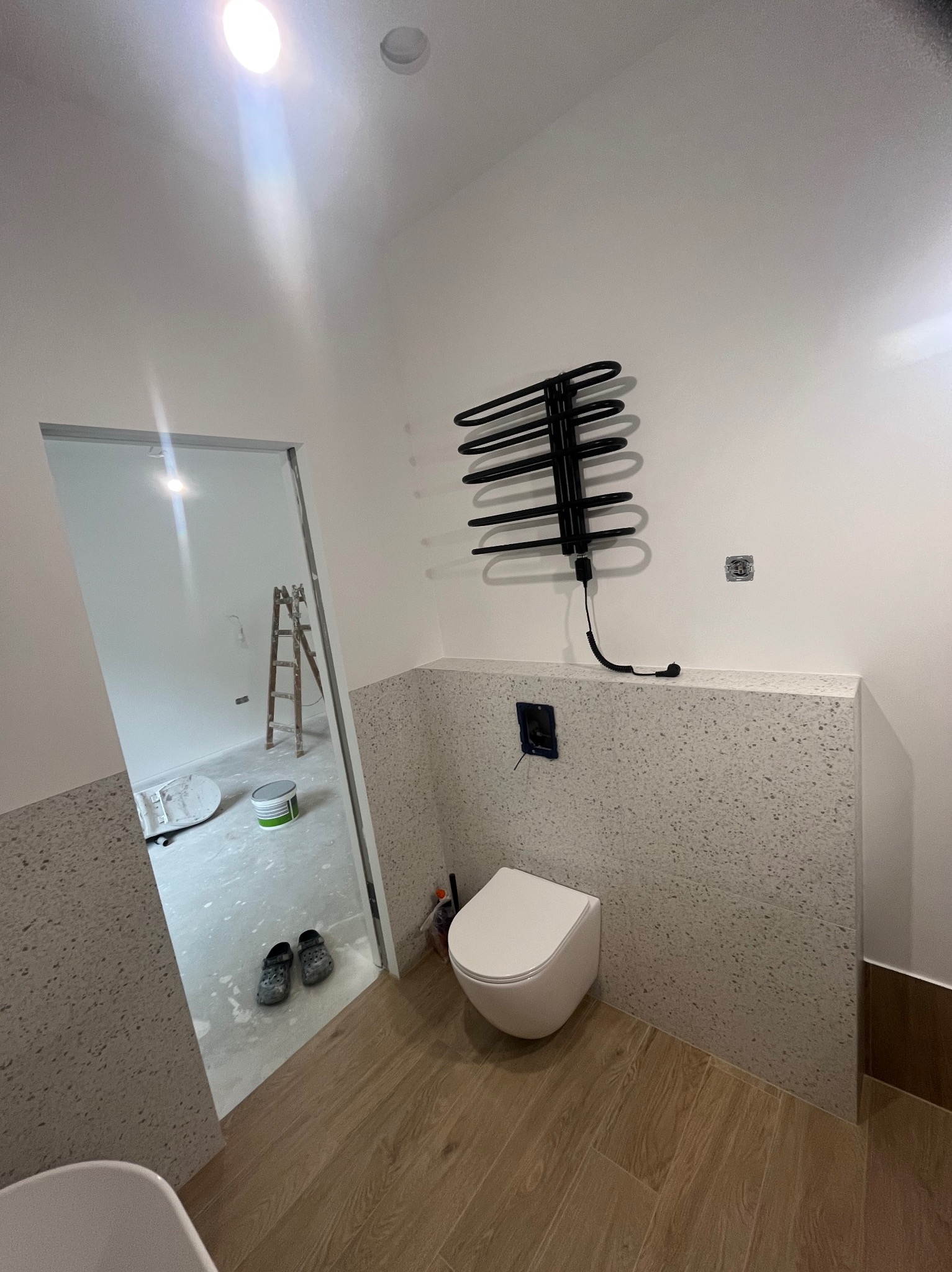 Nowoczesna łazienka w trakcie wykończenia: biała toaleta, grzejnik drabinkowy i ściany wyłożone płytkami w odcieniach szarości. Widoczne prace w sąsiednim pomieszczeniu.