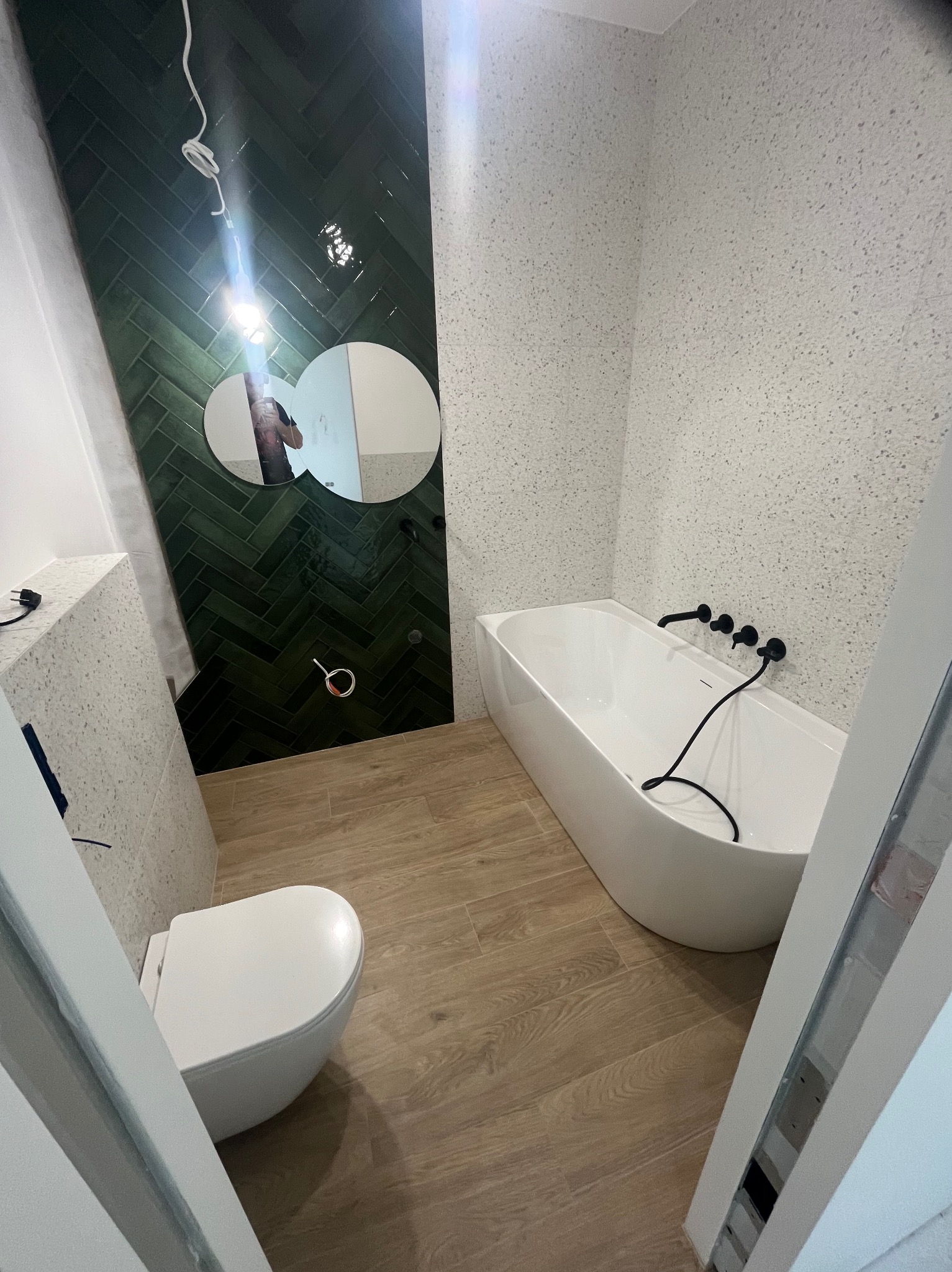 Nowoczesna łazienka po remoncie: zielona ściana z płytek ułożonych w jodełkę, biała wanna z czarną armaturą, drewnopodobne panele i okrągłe lustra.