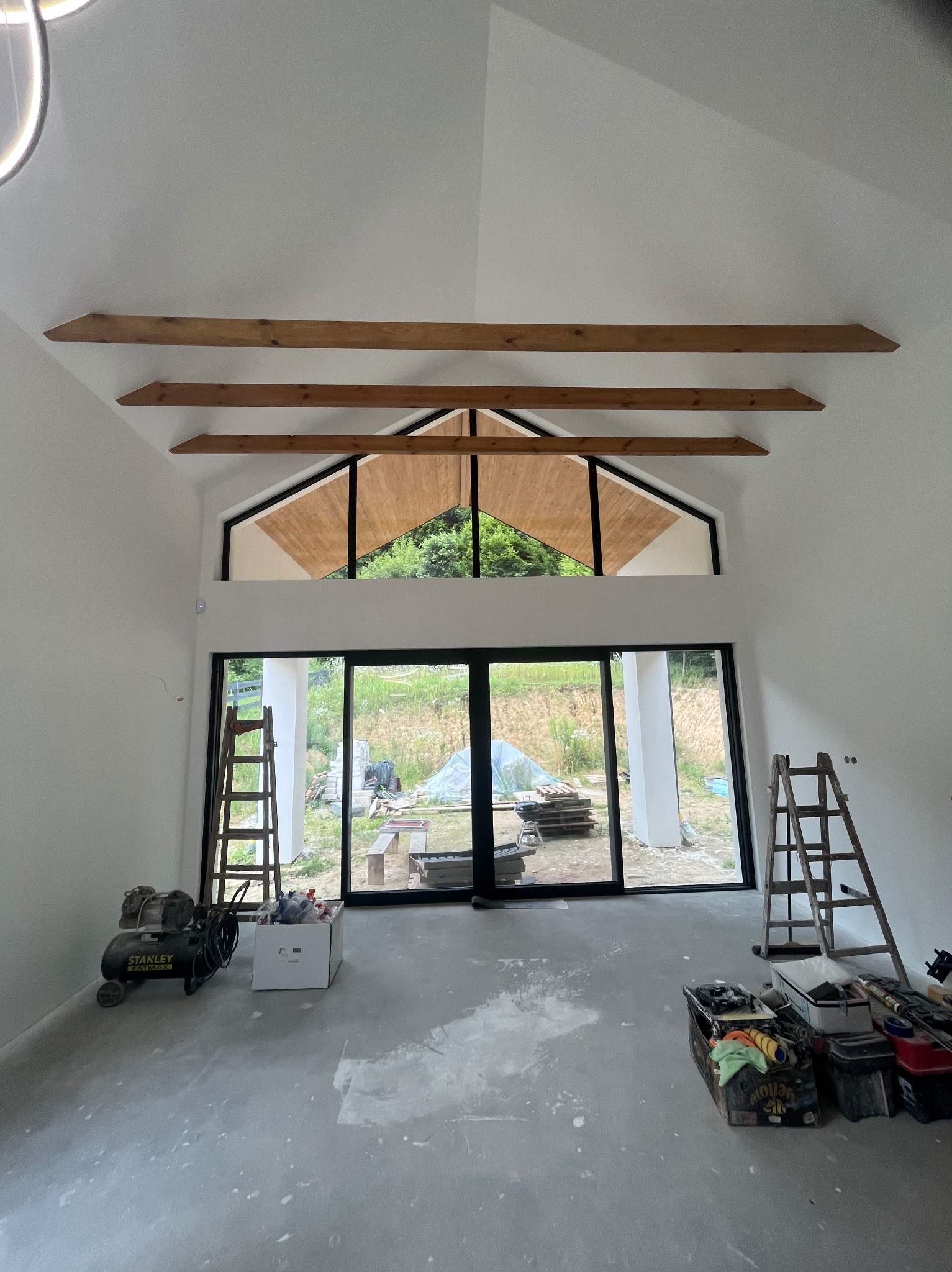 Wysokie wnętrze z drewnianymi belkami i dużym oknem. Widoczne drabiny, kompresor i narzędzia malarskie. Podłoga w trakcie prac wykończeniowych.