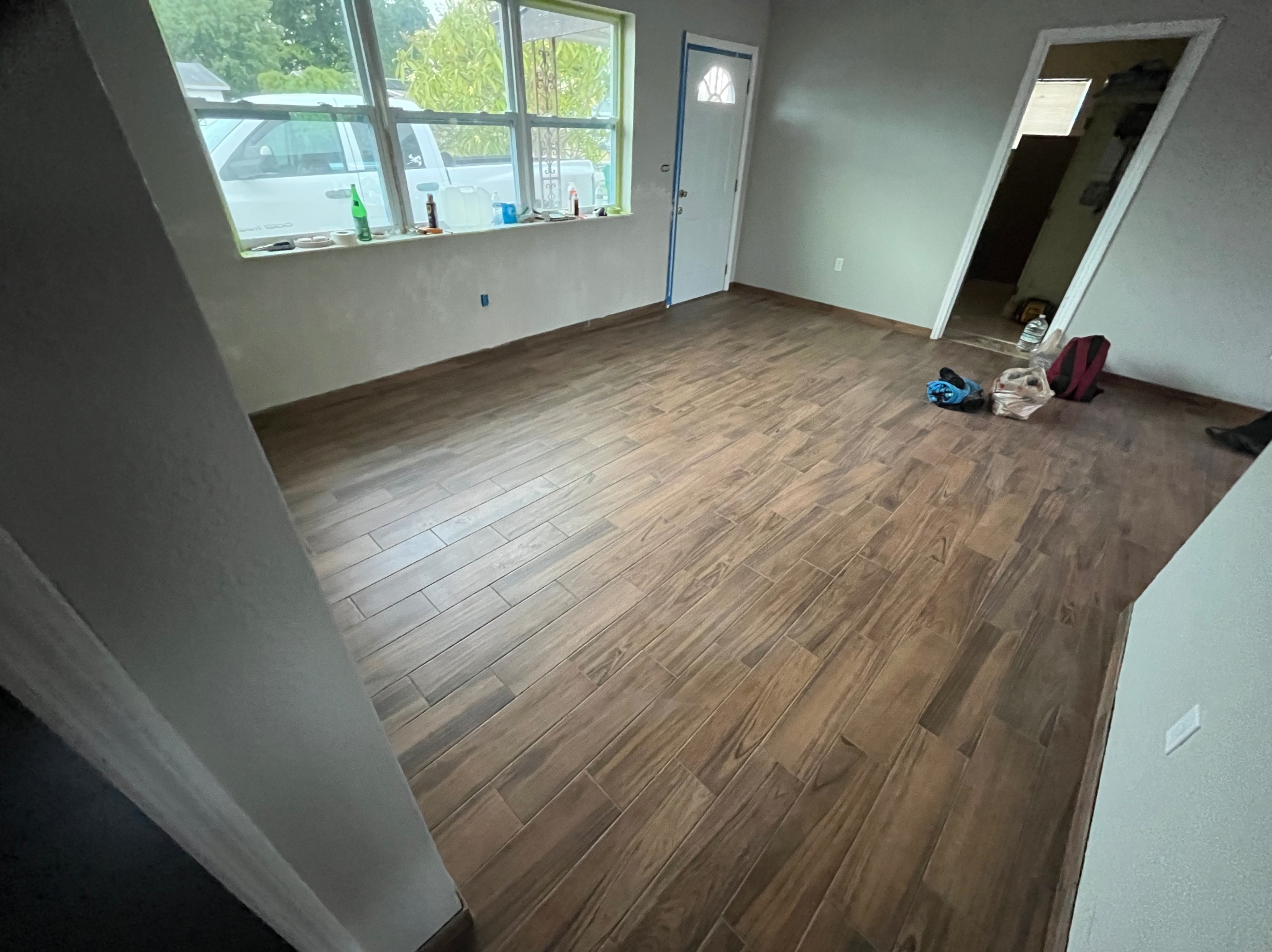 Ułożona podłoga z płytek imitujących drewno w jasnym wnętrzu. Widoczny fragment okna i drzwi z niebieską taśmą malarską. Praca w trakcie wykończenia.