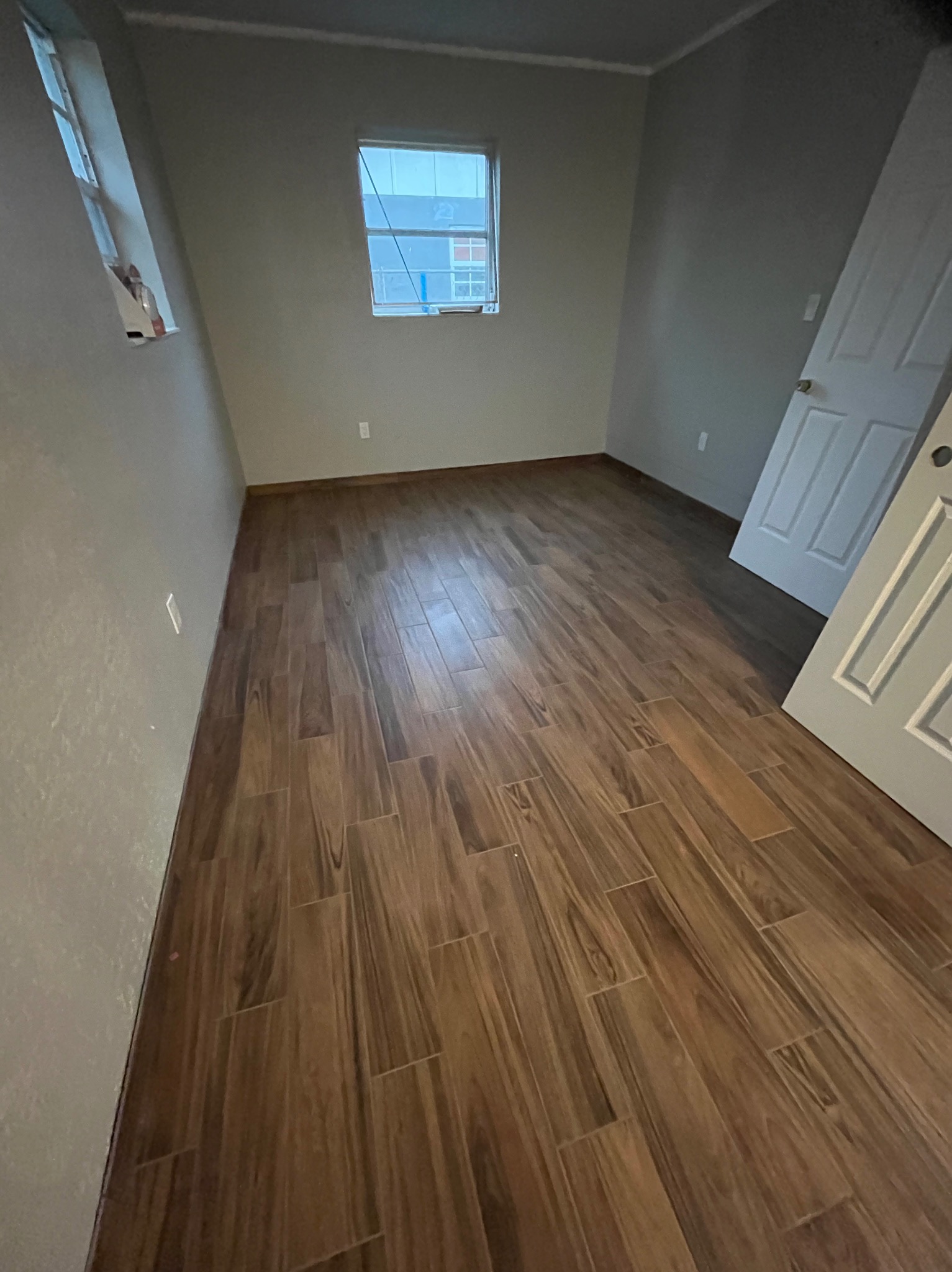 Pokój po remoncie: brązowe panele podłogowe imitujące drewno, szare ściany, białe drzwi i okno. Widoczne gniazdka elektryczne na ścianach.