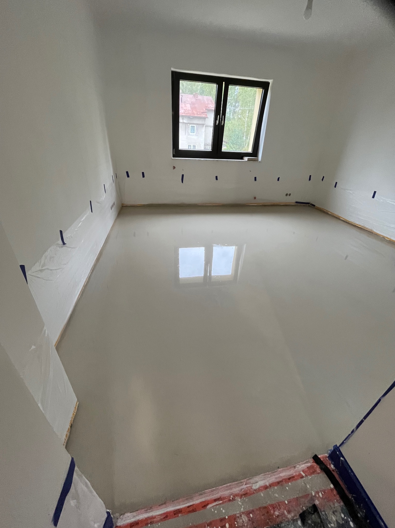 Wylany samopoziomujący podkład podłogowy w jasnym pokoju z oknem. Ściany zabezpieczone folią i taśmą malarską. Widoczne narzędzia malarskie.