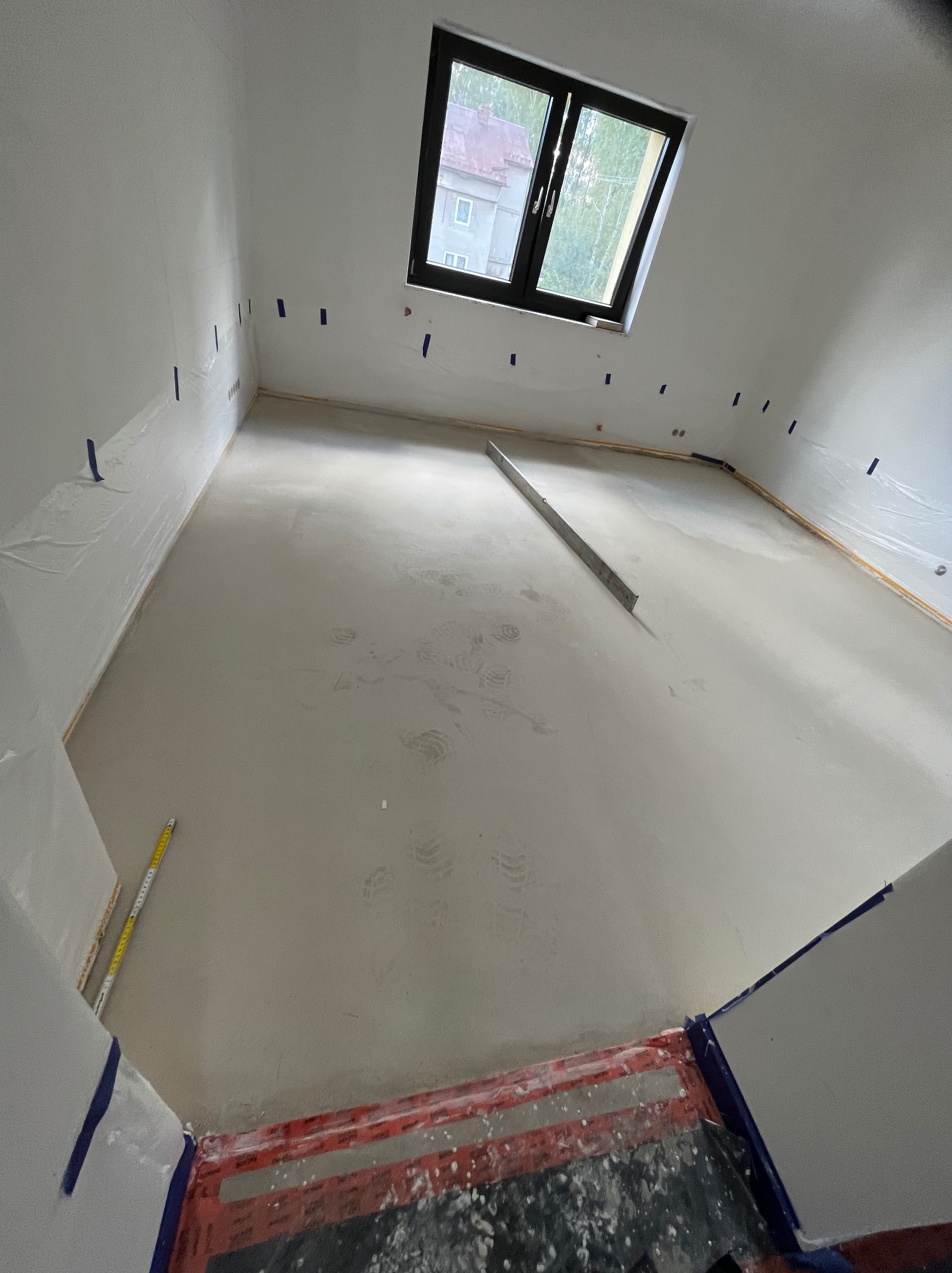Świeżo wylana, jasna posadzka w pokoju z oknem dachowym. Ściany zabezpieczone folią i taśmą malarską. Na podłodze widoczne ślady stóp i długa poziomnica.