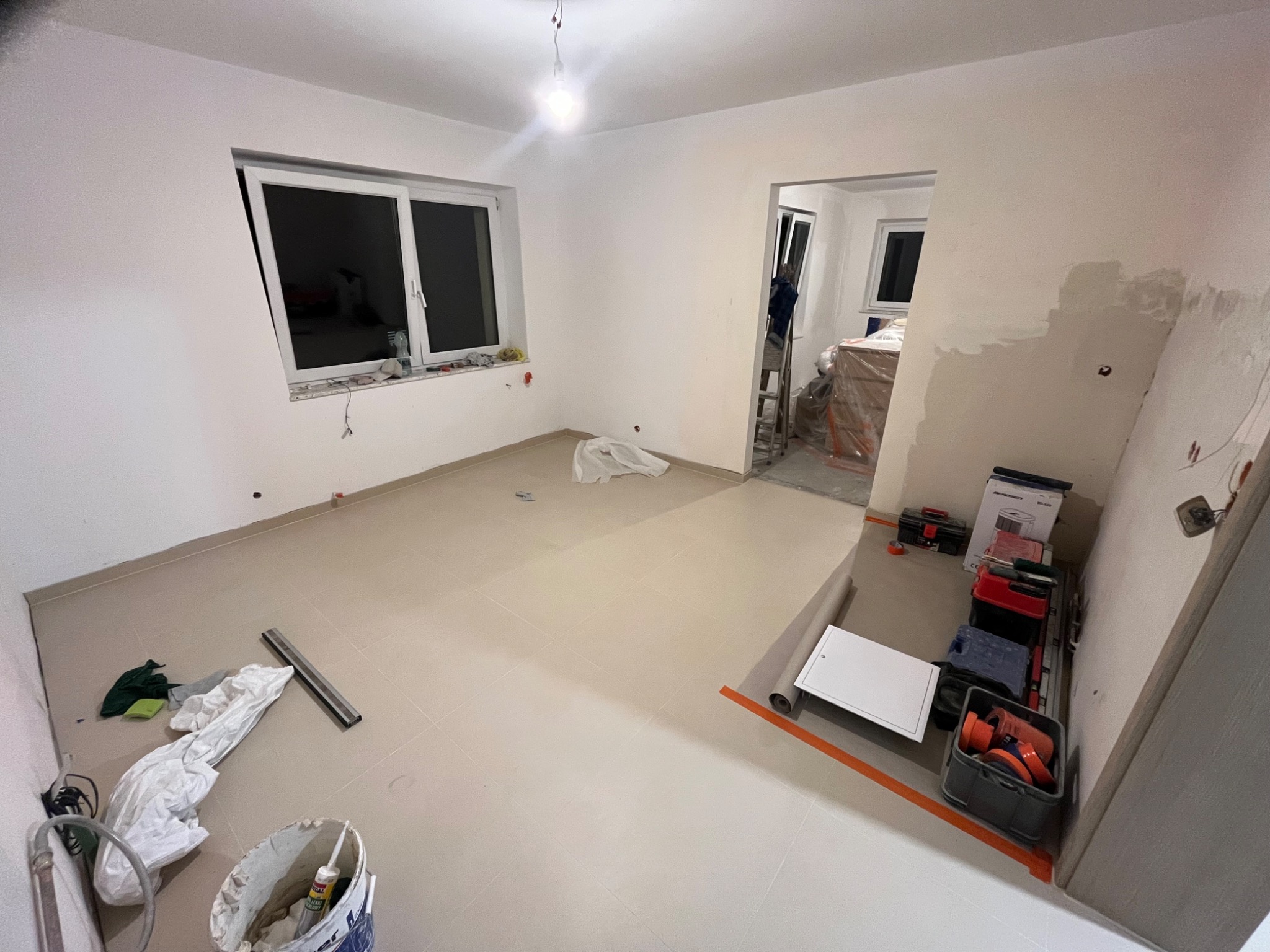 Pokój w trakcie prac wykończeniowych: położona jasna podłoga, częściowo wyszpachlowana ściana, widoczne narzędzia i materiały budowlane. Widok na kolejne pomieszczenie.