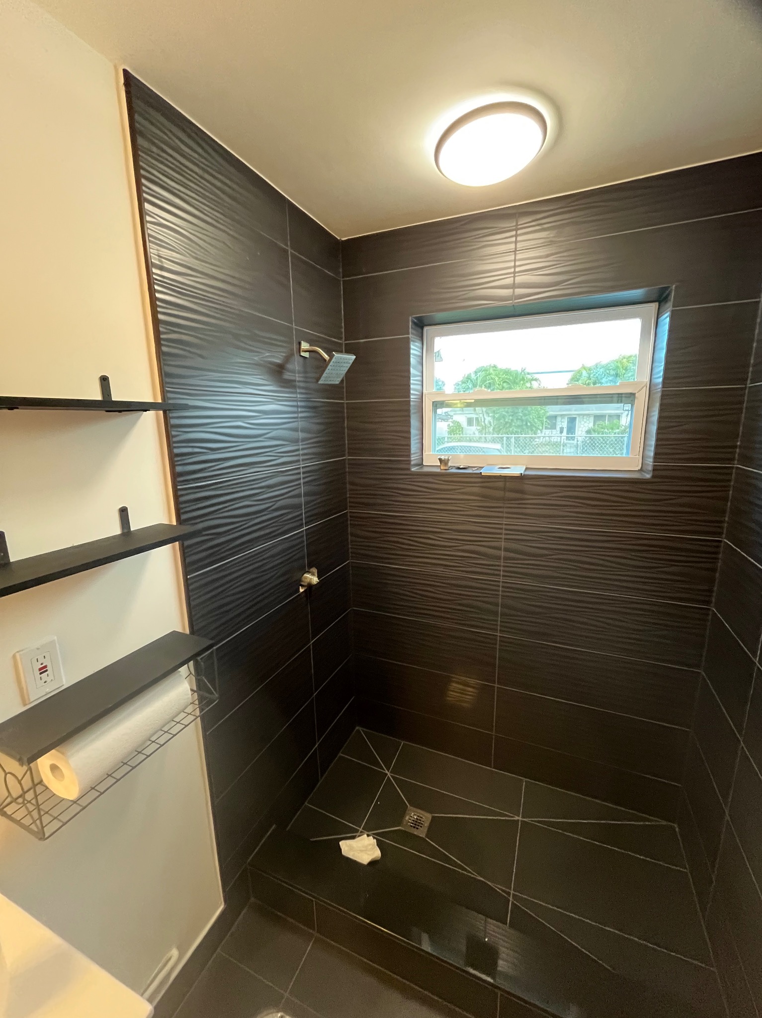 Nowoczesna kabina prysznicowa z czarnymi, falowanymi płytkami, oświetleniem sufitowym i oknem z widokiem na zieleń. Minimalistyczne półki z akcesoriami po lewej stronie.