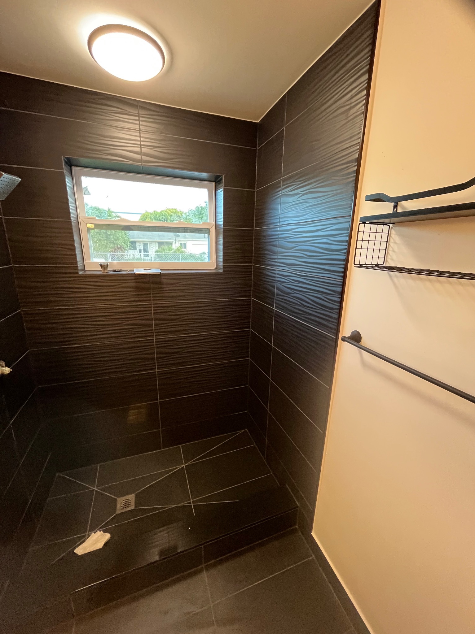 Nowoczesna kabina prysznicowa z ciemnymi, teksturowanymi płytkami i oknem. Na ścianie jasna farba i czarne metalowe półki. Widoczny odpływ liniowy.